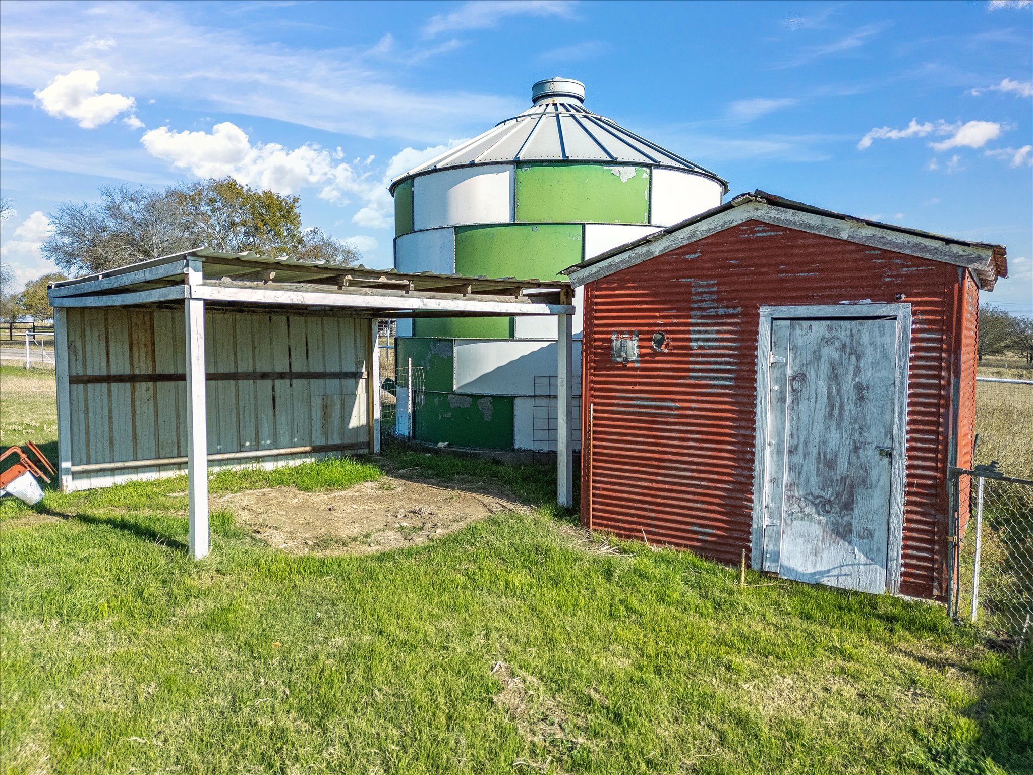 A farm scene with a large green and white silo, a red barn with weathered paint, and a smaller shed with a metal roof, all set against a bright blue sky with scattered clouds and trees in the background.