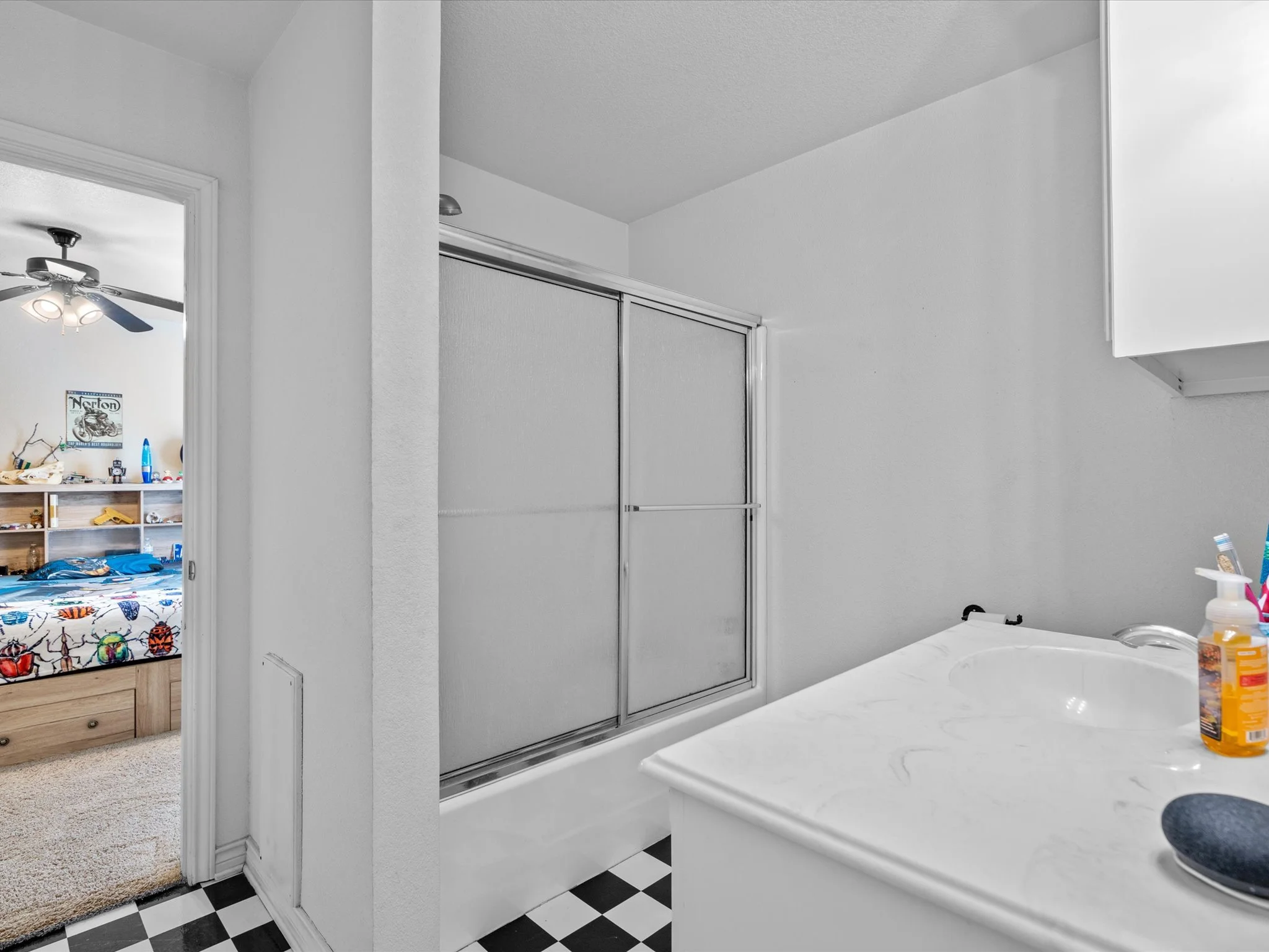 A bathroom with a white counter, a sink with bottles, a sliding glass shower door, and tiled black and white floor. In the adjacent room, a ceiling fan with lights and a cluttered desk are visible.