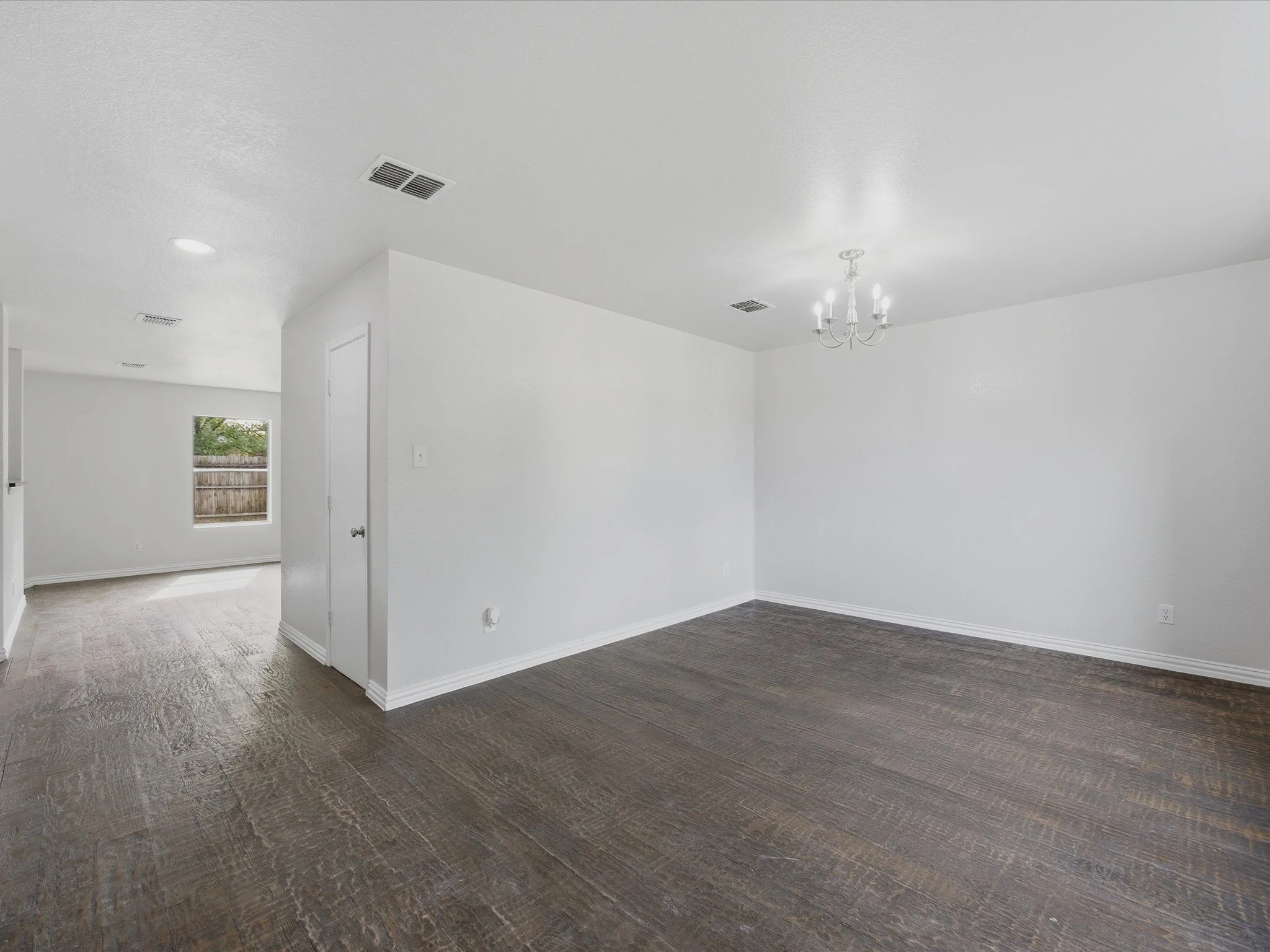 Interior of a white room with dark hardwood flooring, a hanging chandelier, window, and a door.