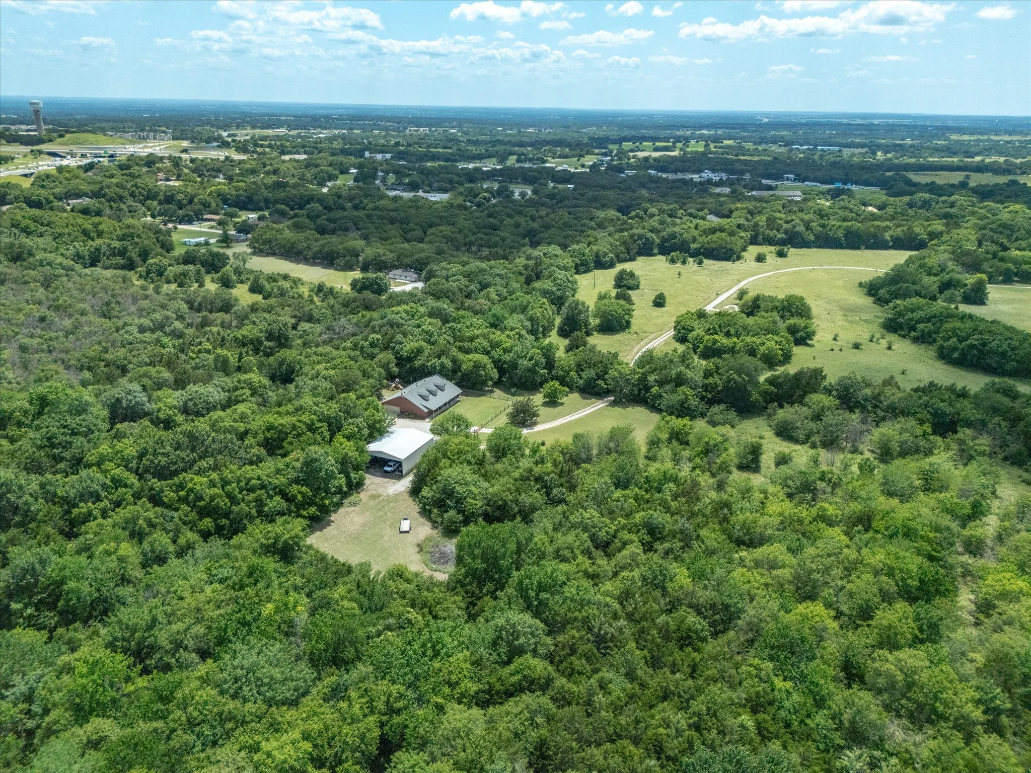 Aerial view of a green landscape with trees, houses, a road, and a distant highway under a blue sky with scattered clouds.
