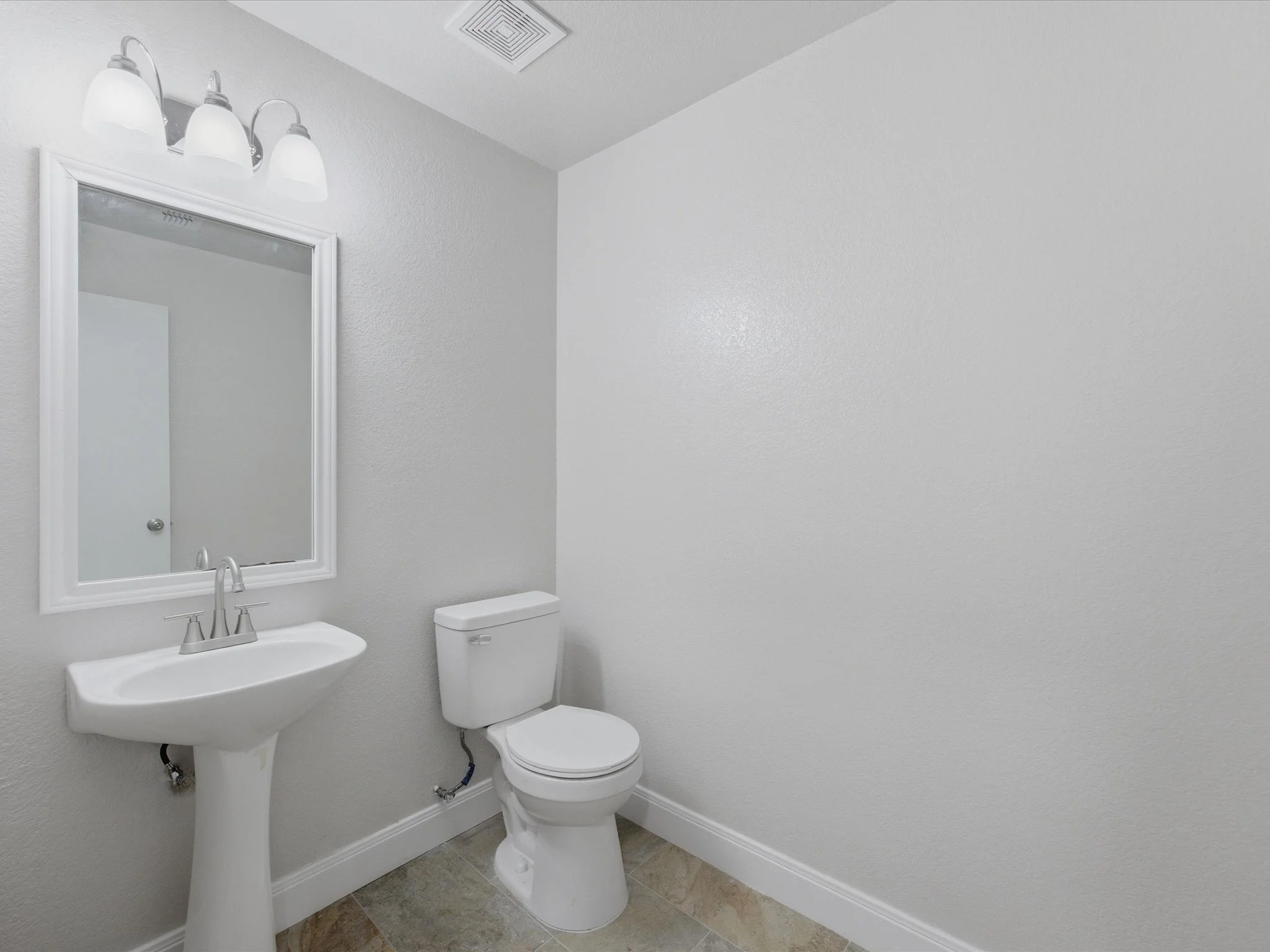 A small bathroom with a pedestal sink, a toilet, a mirror with a white frame, and a three-light vanity fixture. The walls are painted light gray and there is a ceiling vent.