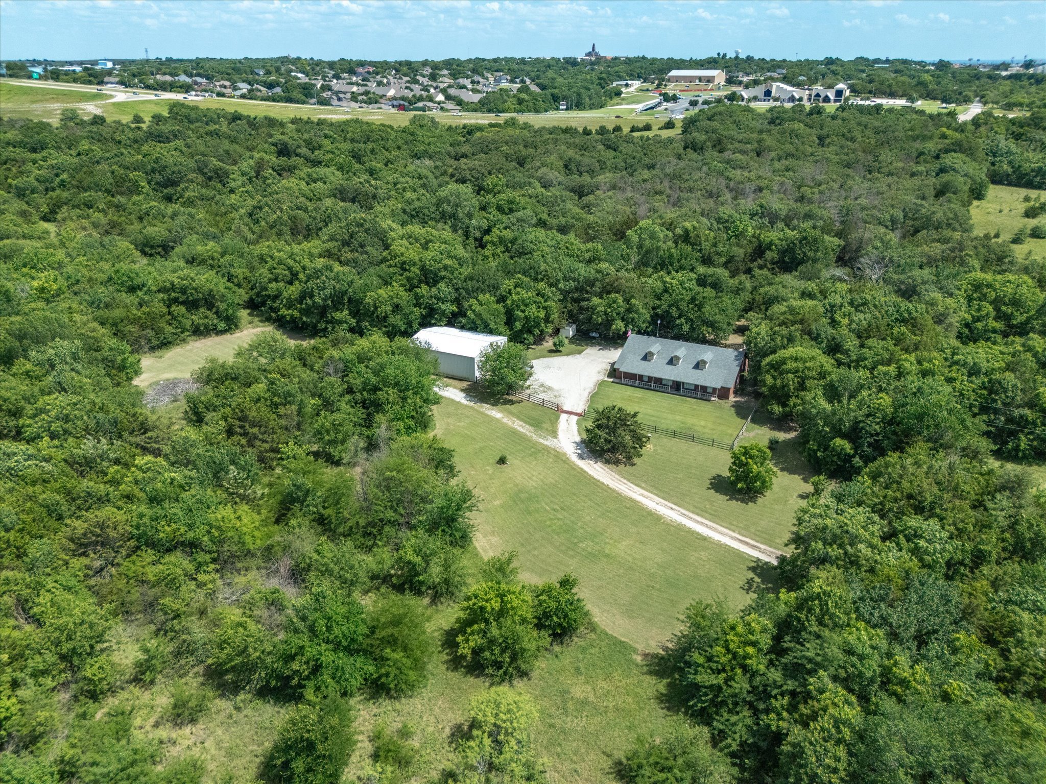 An aerial view of a rural property with a house, a metal building, and fencing surrounded by dense trees, with a wooded area extending toward a suburban neighborhood in the background.