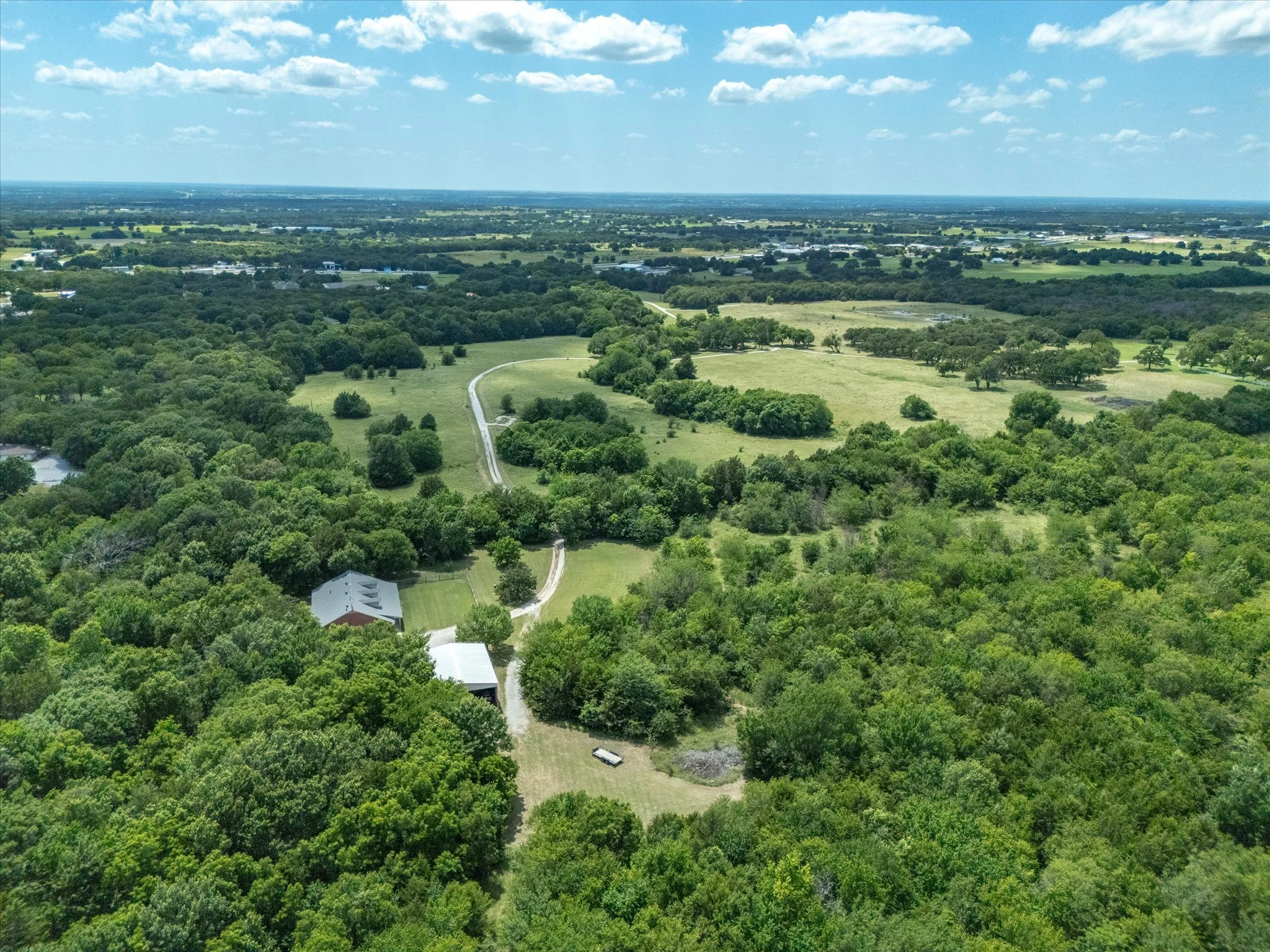 Aerial view of a rural landscape with green trees, open fields, a winding road, and a few buildings under a partly cloudy sky.
