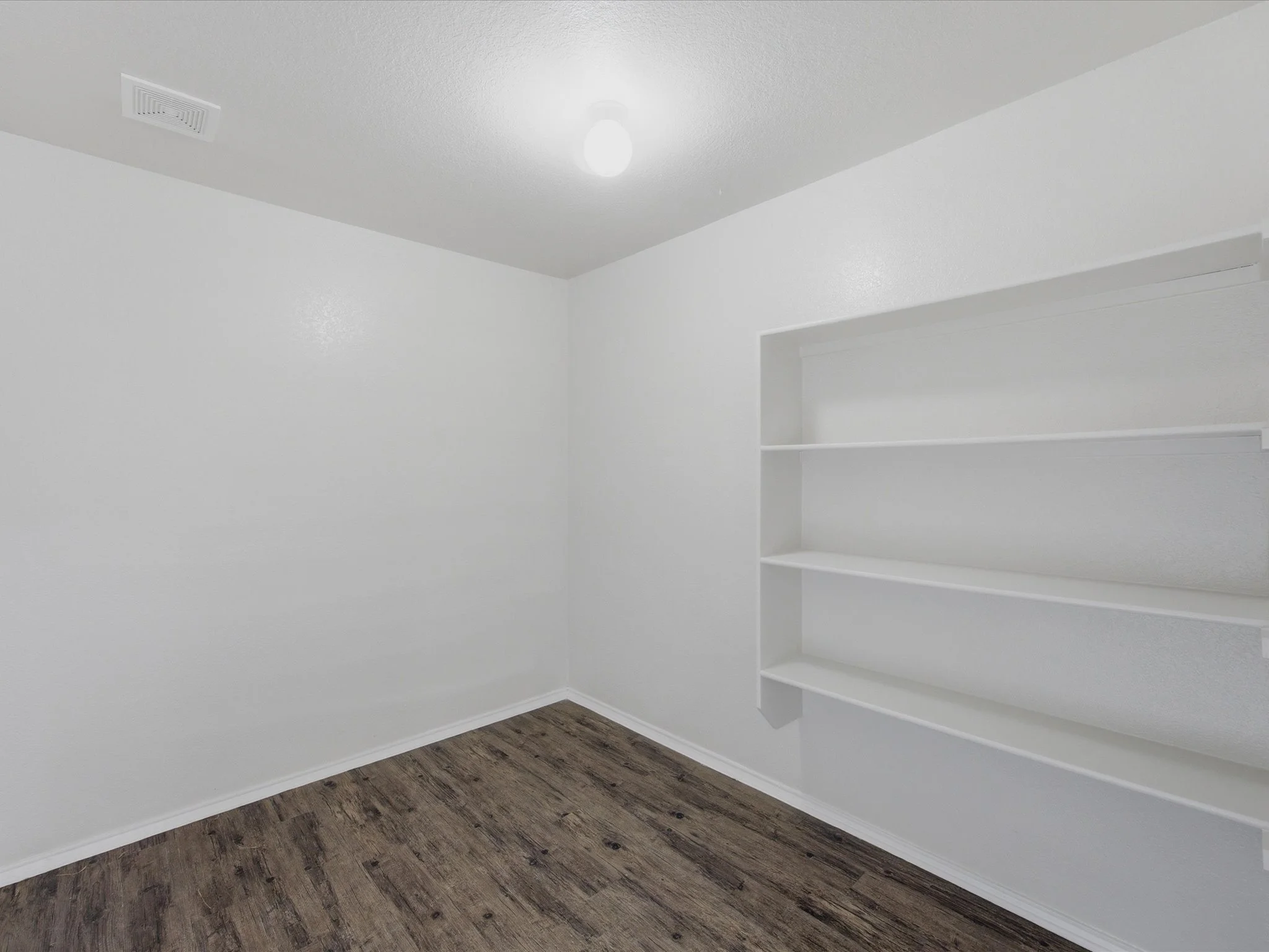 A white interior room with a white ceiling, a ceiling vent, a lightbulb, a corner with white shelves, white walls, and wood-look flooring.