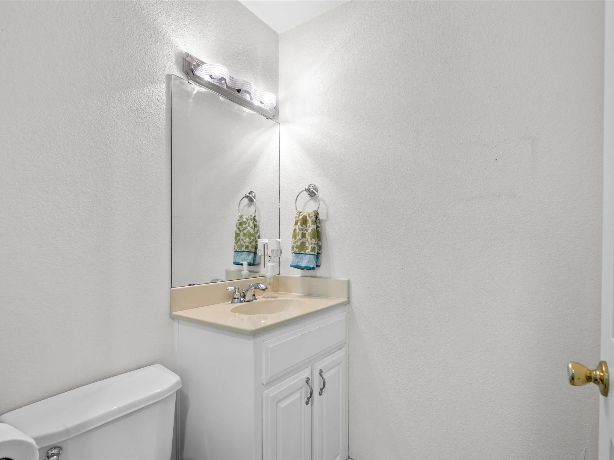 A bathroom sink with a beige countertop, a mirror above it, and two small towels hanging on hooks. The light fixture above the mirror is turned on, casting light on the textured white walls. A white cabinet with two doors is below the sink, and a sma