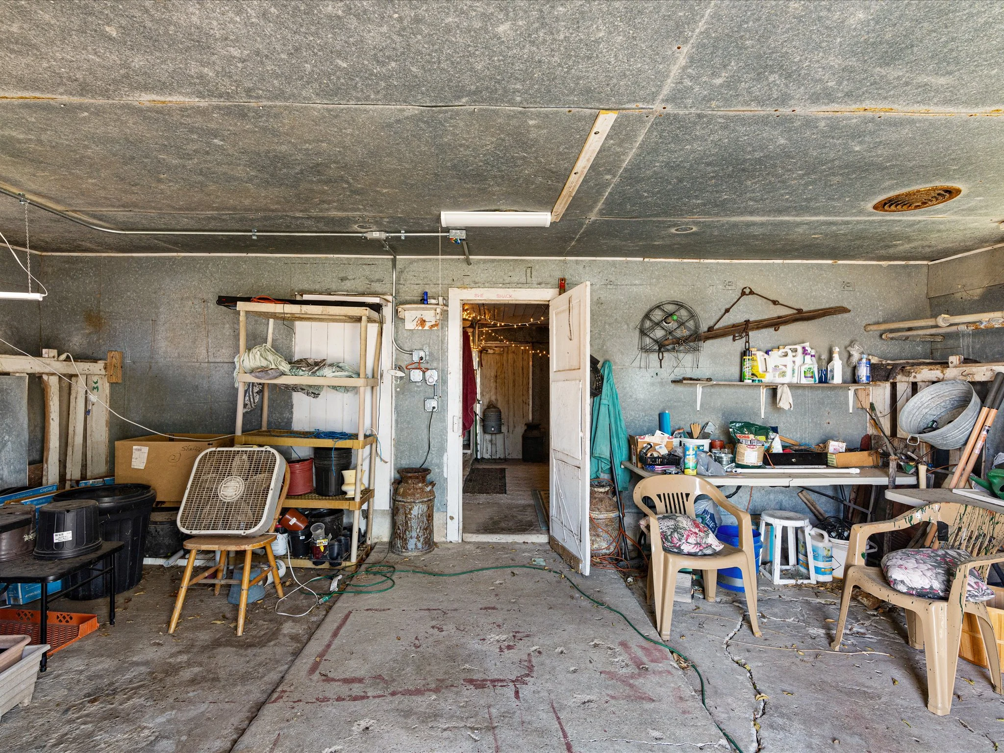 The image shows a cluttered garage with various tools, chairs, and storage items scattered around. The walls are gray with some hanging items, and the ceiling is unfinished with visible pipes and a light fixture. An open doorway leads to another room