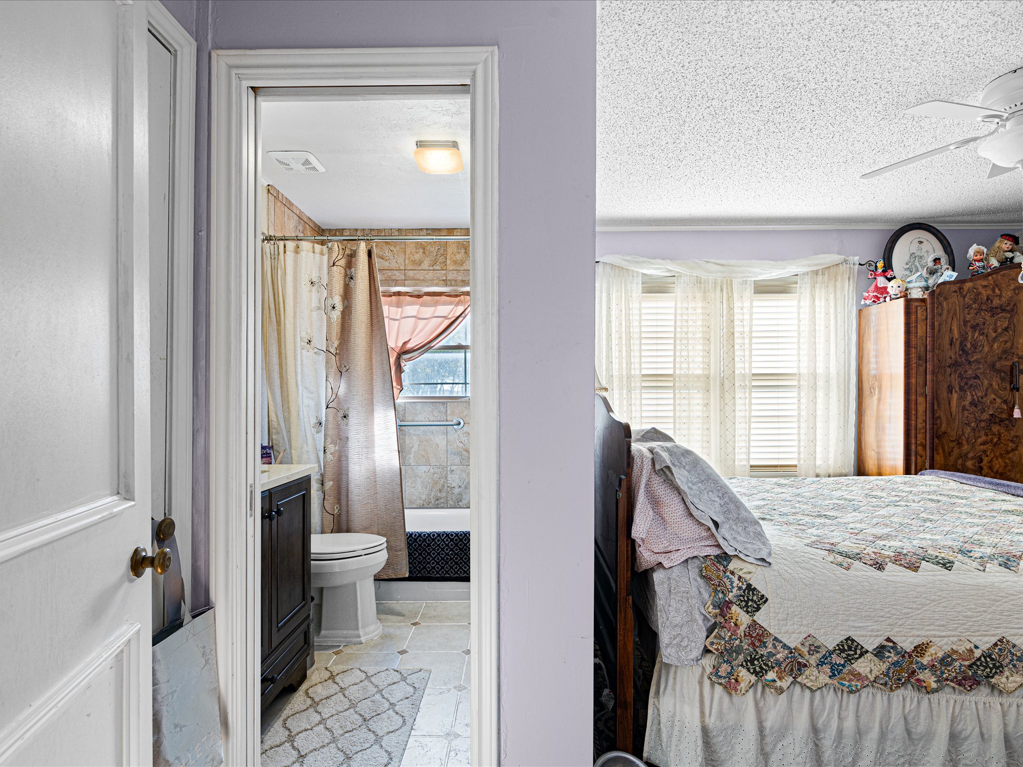 View of a bedroom and bathroom from the doorway showing a bed with a colorful quilt, a dresser with stuffed animals and decorations, and a bathroom with a toilet, shower curtain, and window with curtains.
