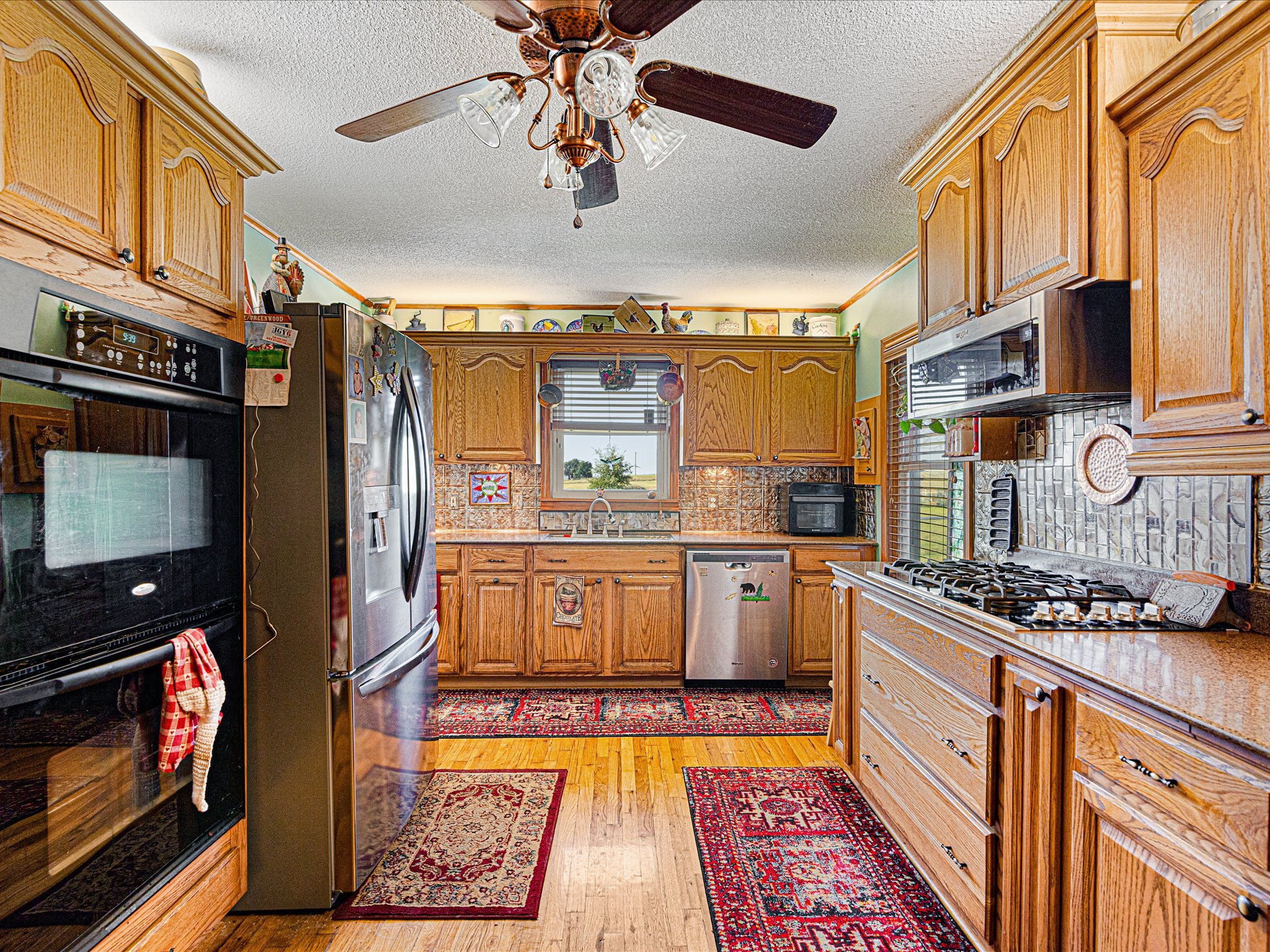Kitchen with wooden cabinets, stainless steel refrigerator, oven, microwave, and colorful decorations, including a patterned rug and window with view of outdoor landscape.