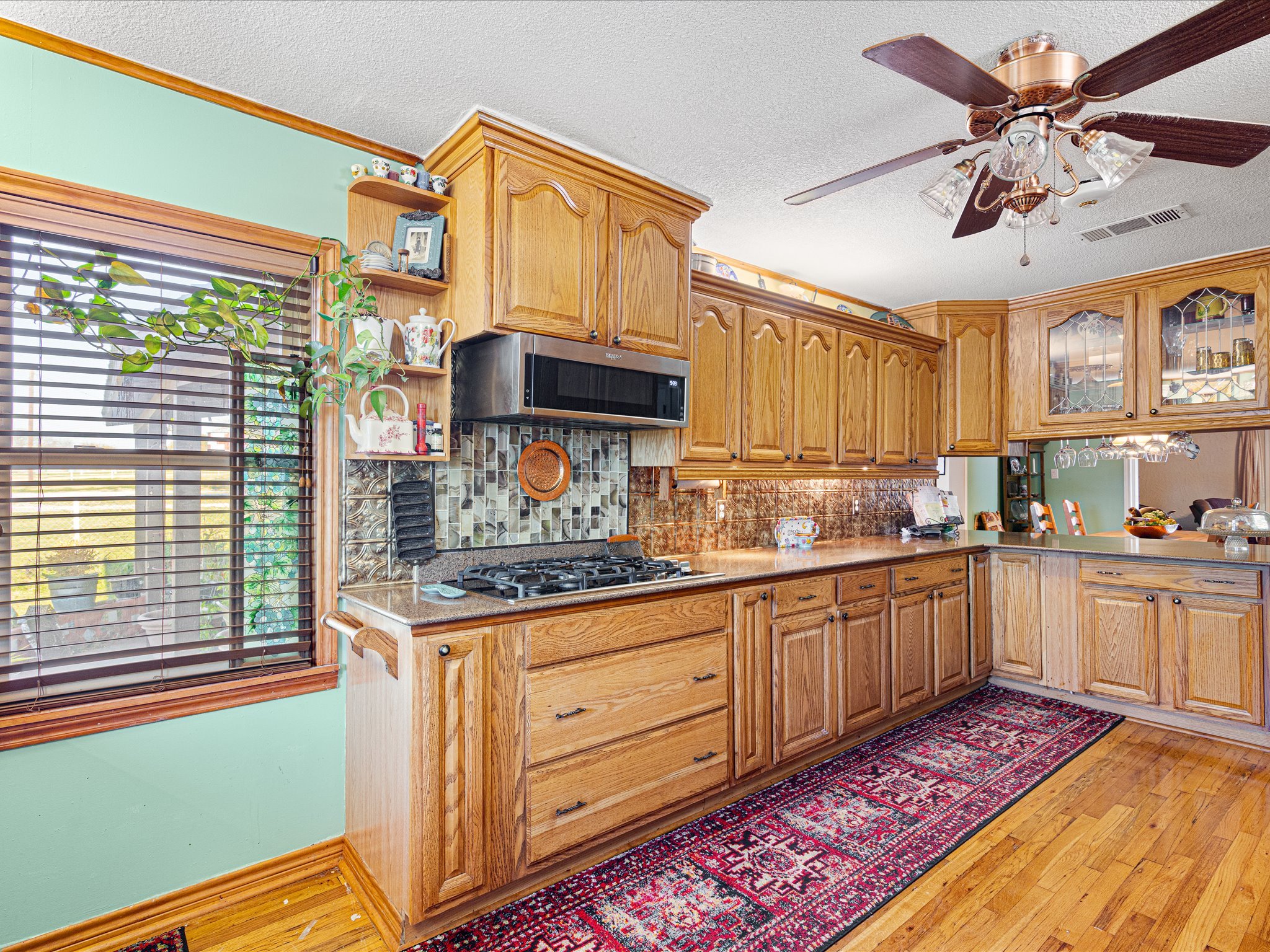 A kitchen with oak cabinets, a countertop with a colorful patterned runner rug, a microwave oven, a gas stove, a plant near a window with blinds, and a ceiling fan with lights.