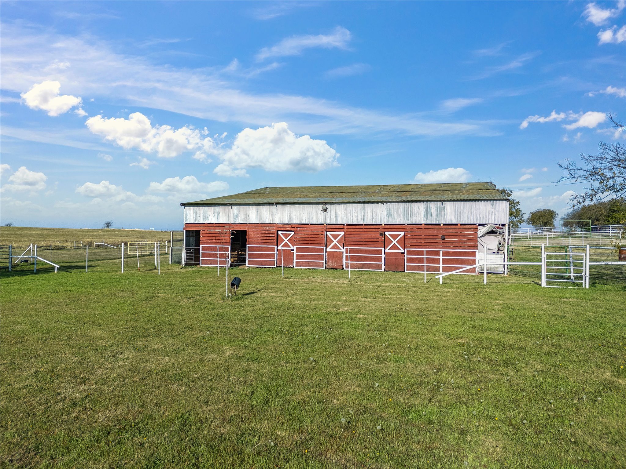 A red barn with a weathered metal roof and white trim, surrounded by green grass and a white fence, under a partly cloudy sky.
