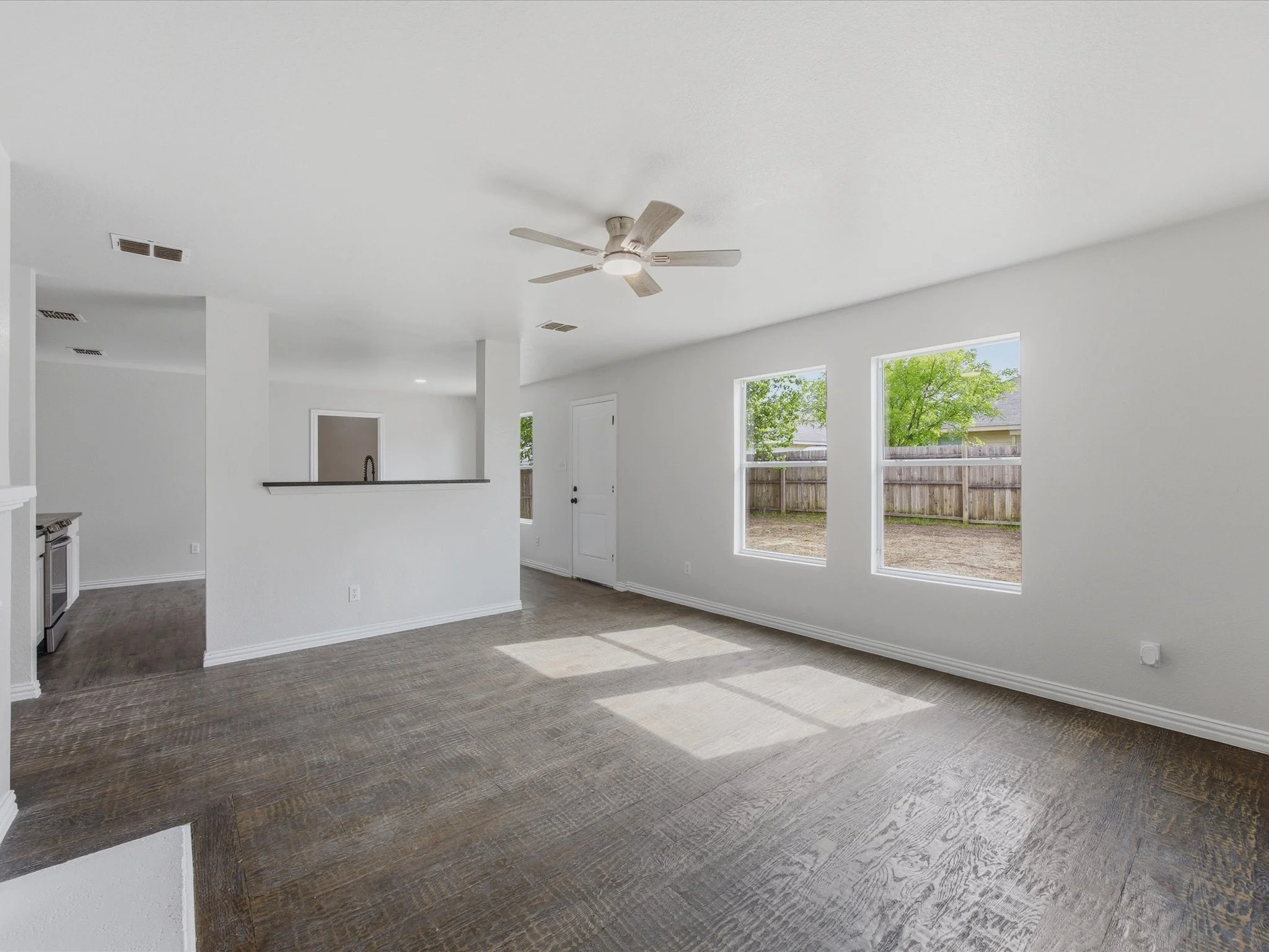 Bright living room with large windows, gray carpet, white walls, ceiling fan, and outdoor yard visible.