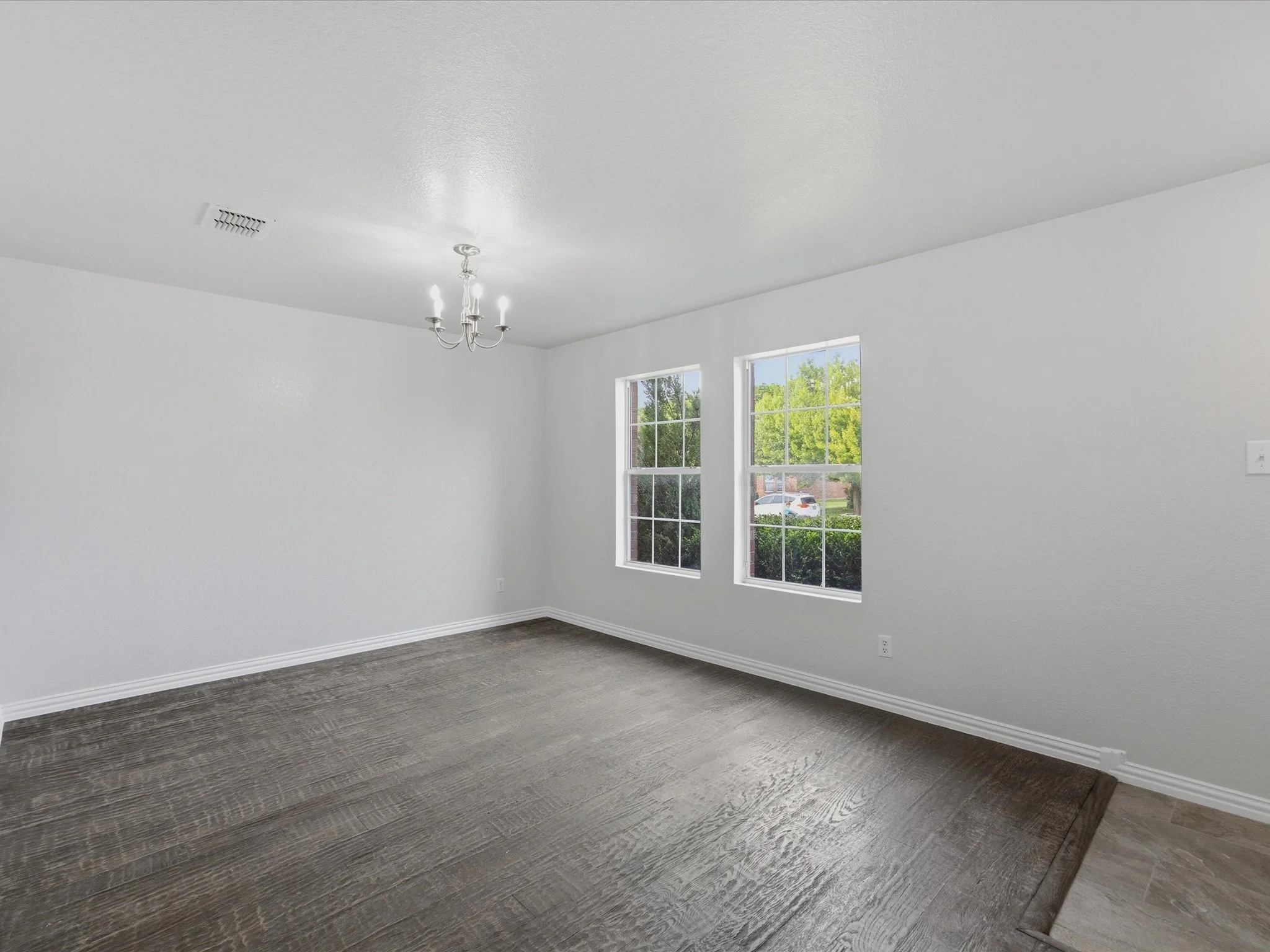 Empty living room with large windows, white walls, dark wood flooring, a chandelier, and a ceiling vent.
