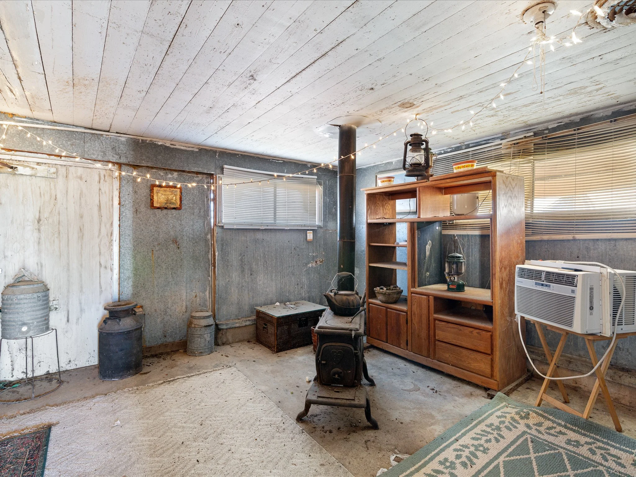 Interior of an old, rustic room with a white, worn wooden ceiling, string lights, a wood-burning stove, metal containers, an air conditioner, a small shelf with bowls and lamps, and a window with blinds.
