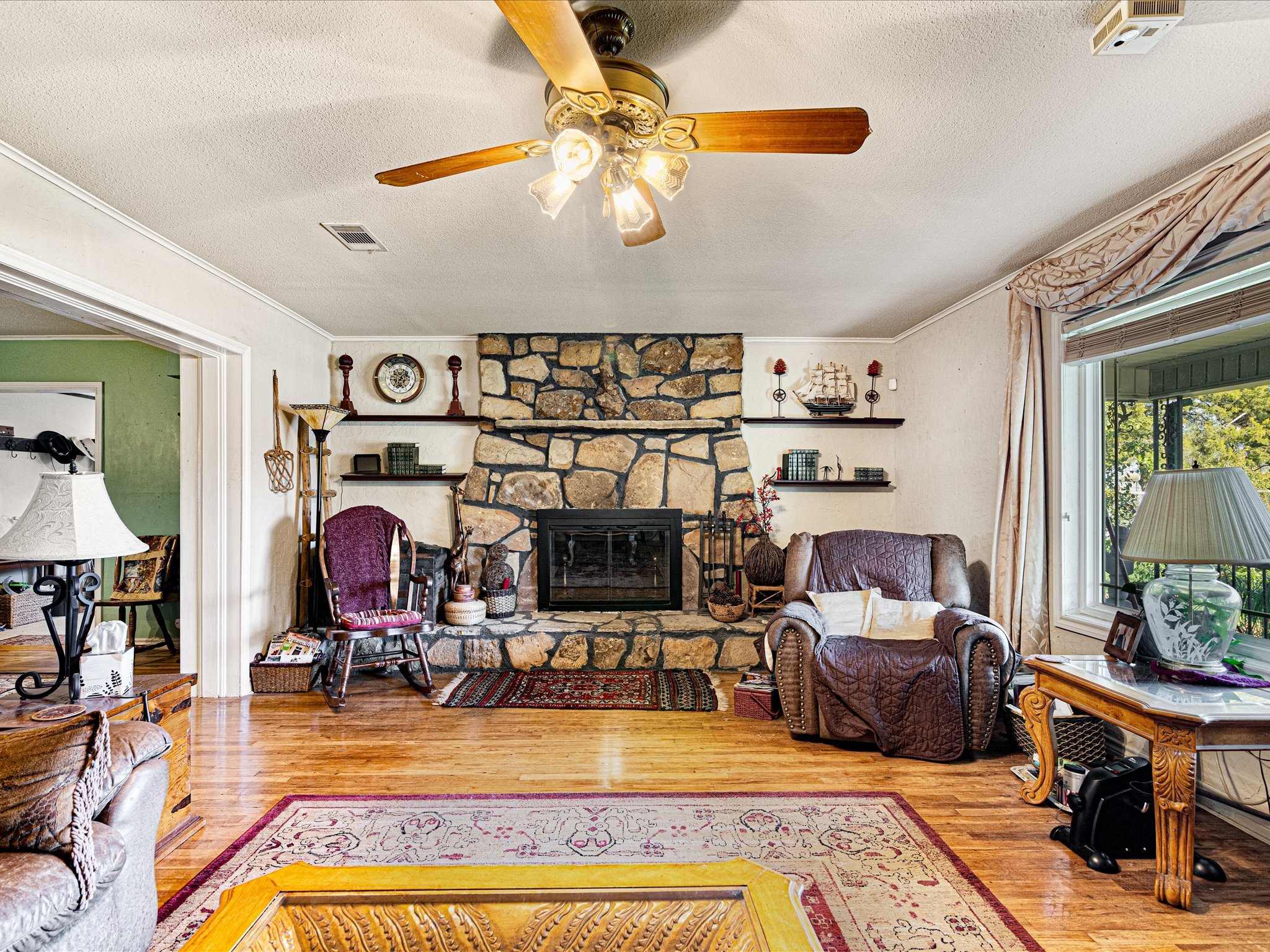 Living room with a stone fireplace, wooden floor, and various furniture and decorations, including armchairs, shelves with books and decorative items, a ceiling fan with lights, window with curtains, and a view of outdoor greenery.