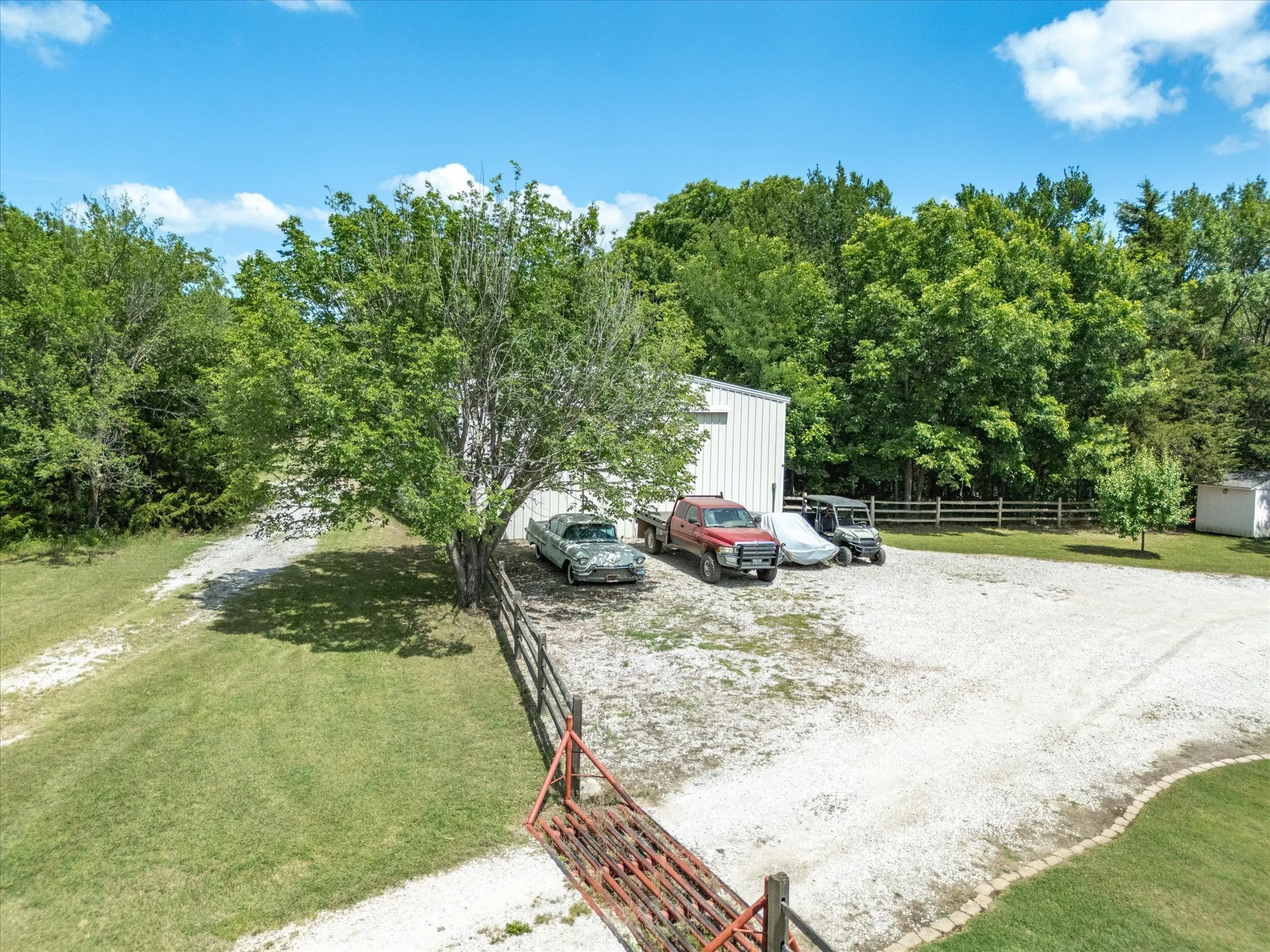 A gravel driveway with several old cars parked near a large white building, surrounded by green trees and a blue sky with some clouds.