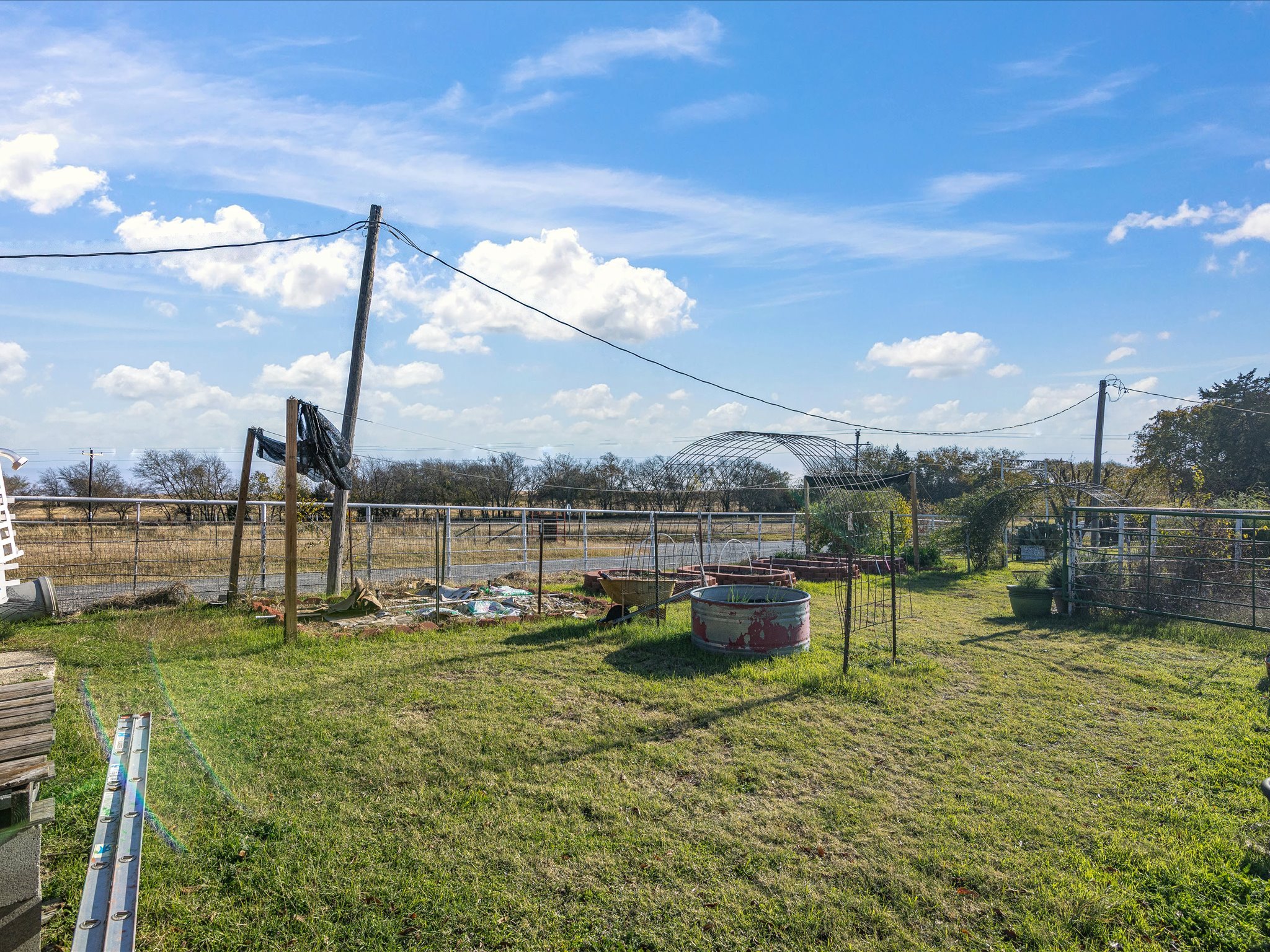 A rural backyard with a clear blue sky, some scattered clouds, and visible utility poles with wires. The yard has green grass, various garden beds, and some structures, including a wooden frame and fencing.