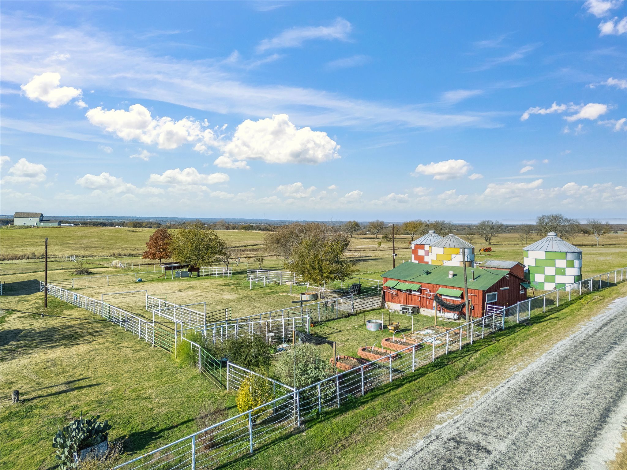 Open rural landscape with green fields, a few trees, farm buildings with colorful round roofs, and a bright blue sky with scattered clouds.