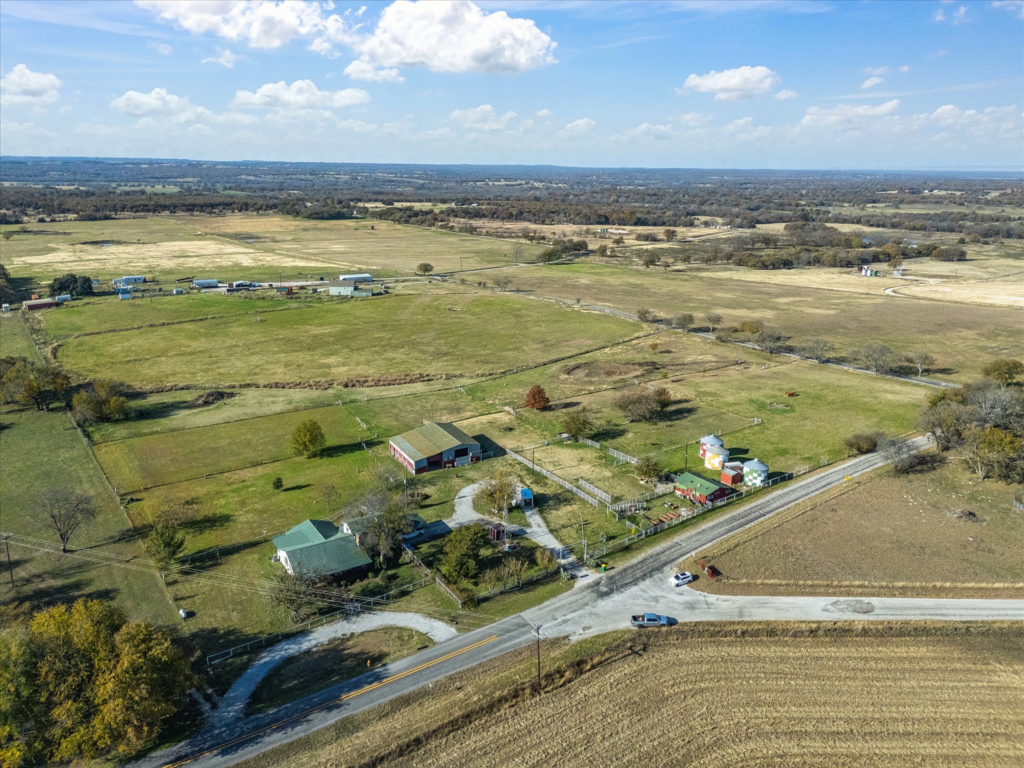 Aerial view of a rural landscape with farmland, a few scattered houses and buildings, a main road with cars, open fields, and distant hills under a partly cloudy sky.