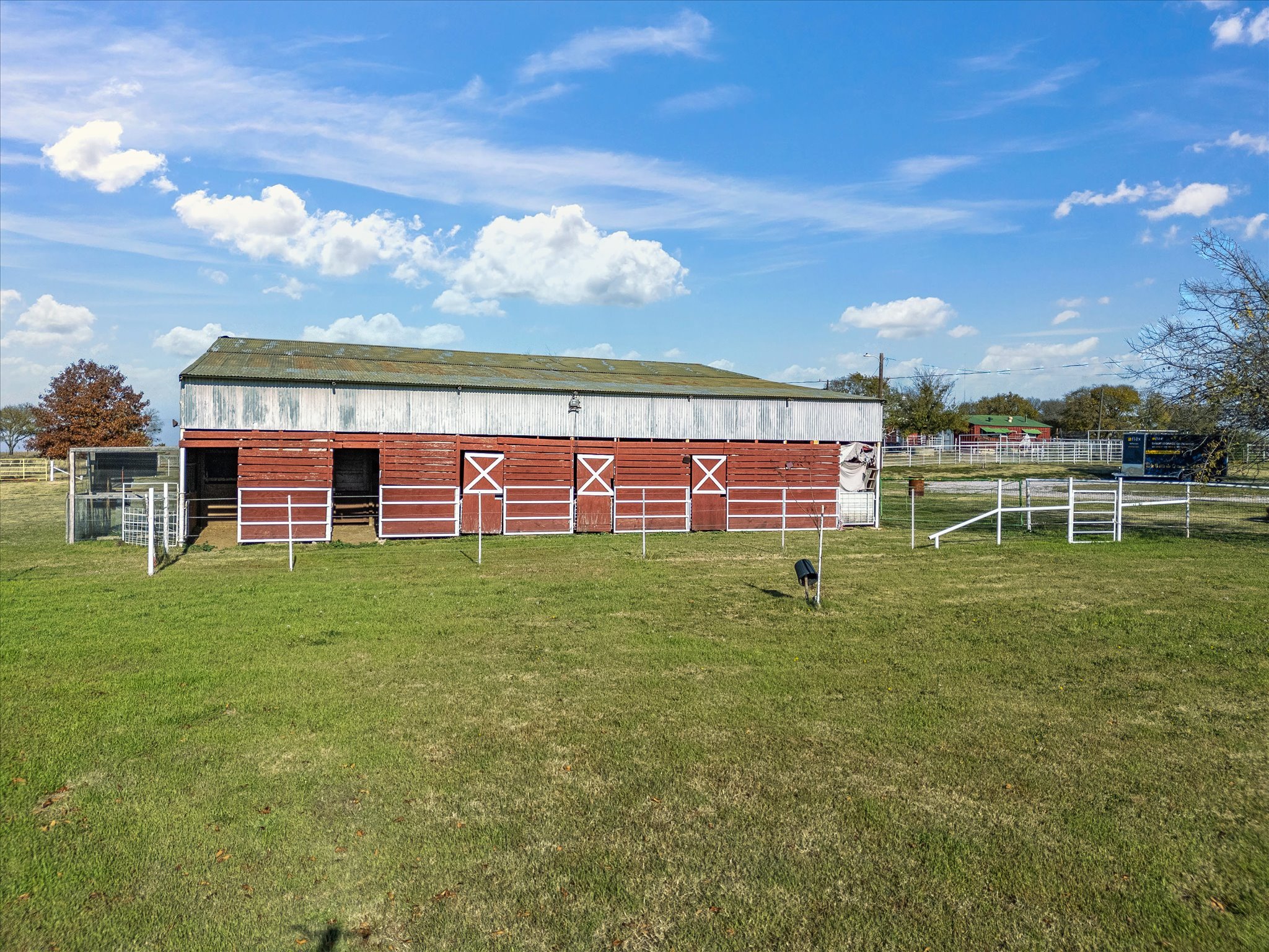 A large red barn with white trim and double doors, surrounded by green grass and fenced areas, under a blue sky with scattered clouds.
