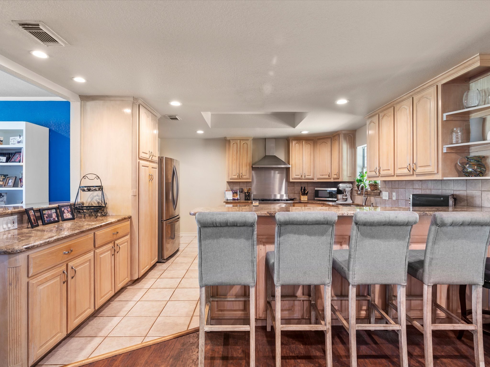 A spacious kitchen with wooden cabinets, stainless steel appliances, and a large island with seating for four. The kitchen has a tiled floor, a blue accent wall, and various decorative items.