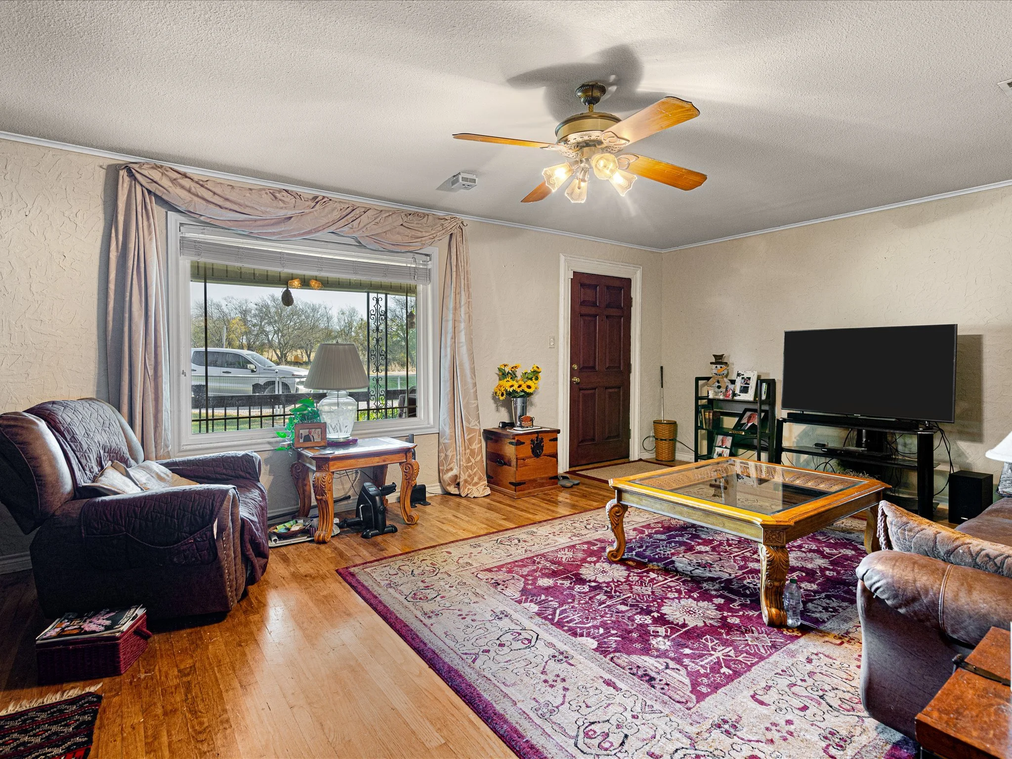 Living room with beige textured walls, hardwood floor, a window with light pink drapes, a dark wooden door, a sofa with pillows, a coffee table, a flat-screen TV, a lamp, a bouquet of yellow flowers, and a decorative snowman.