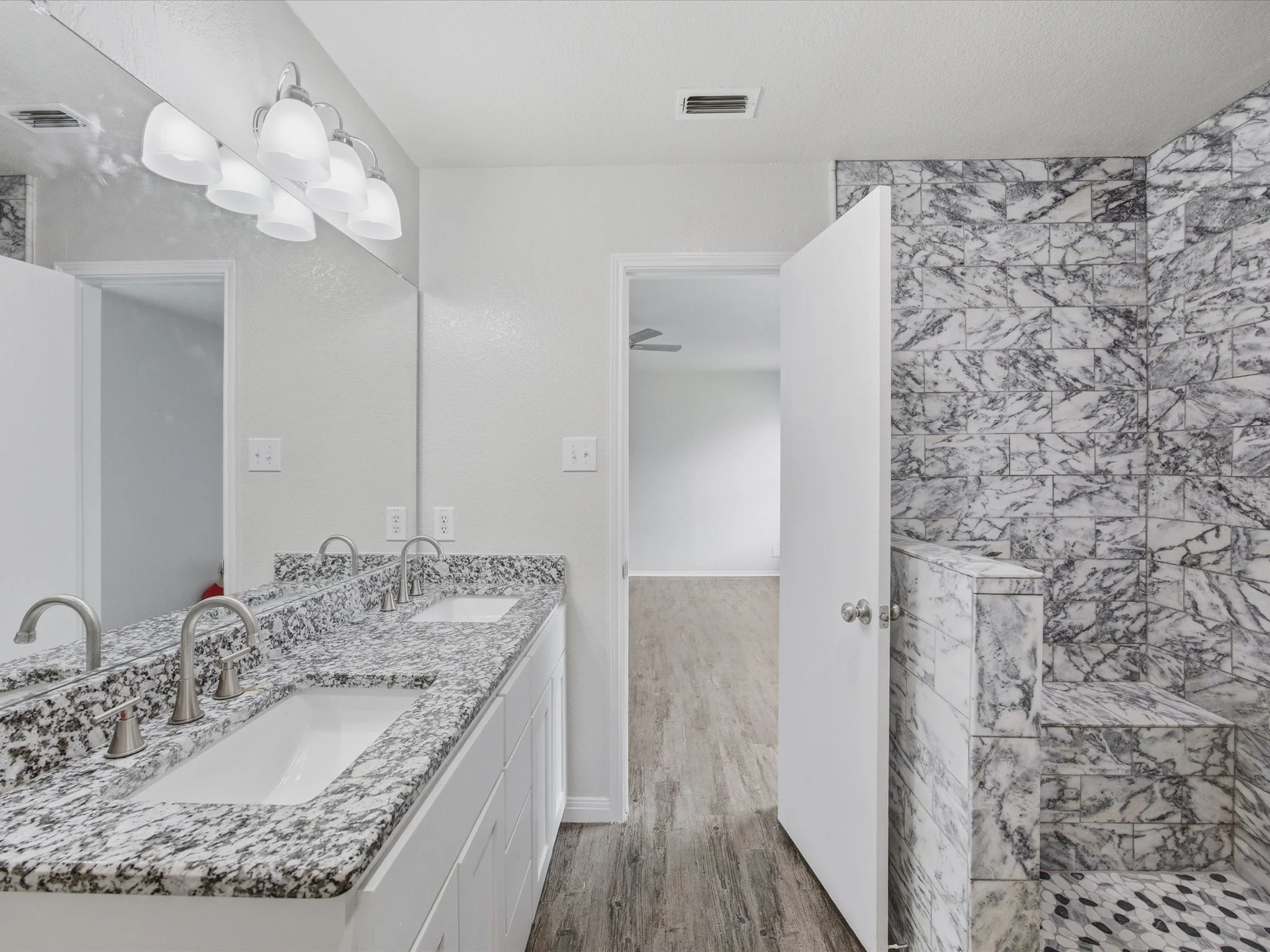 Modern bathroom with white cabinets, granite countertops, a large mirror, and a marble-tiled shower area.