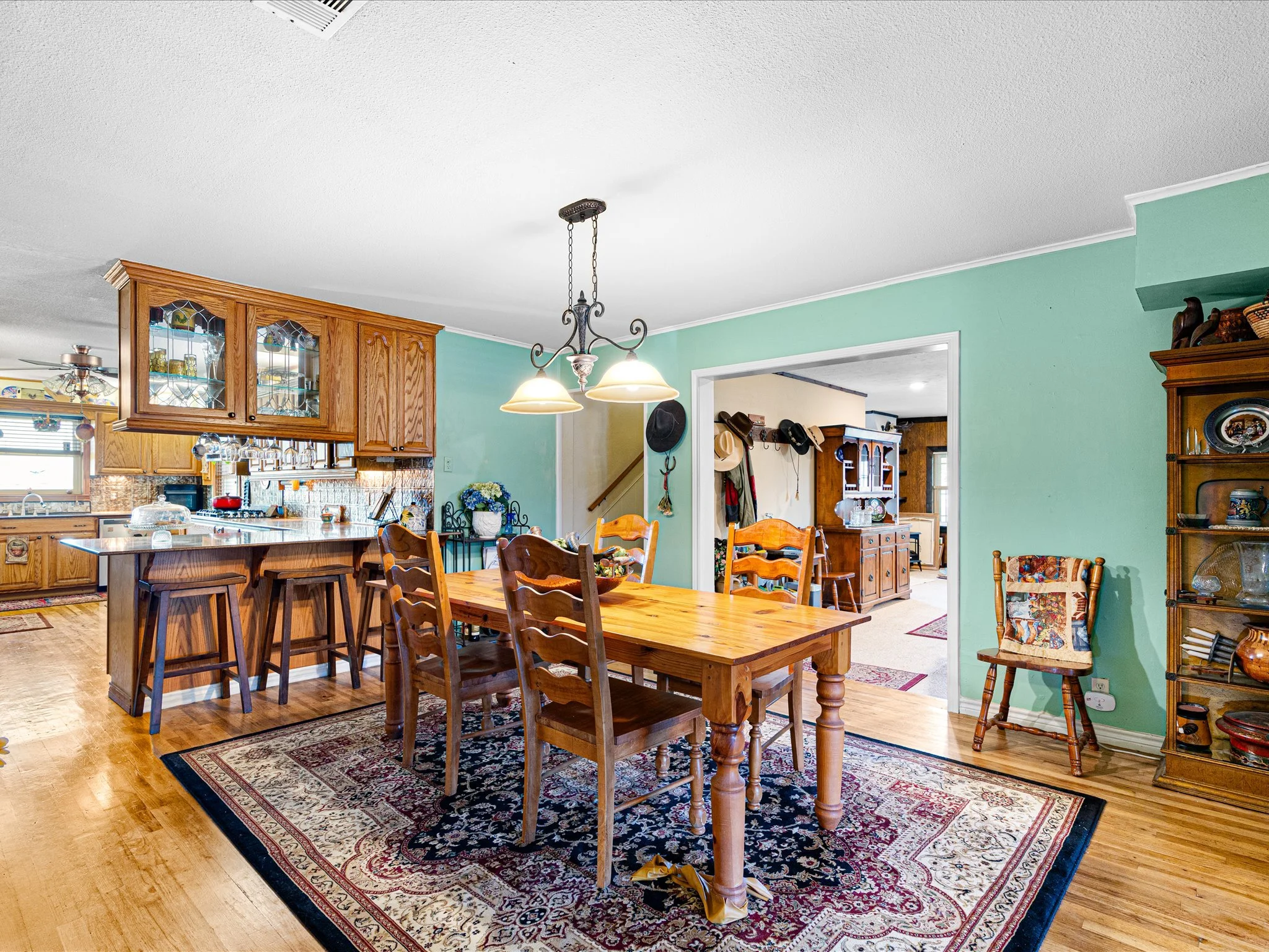 A cozy dining area with a wooden table and matching chairs, a patterned rug, and a green wall. The space features a chandelier with three lamps, a glass-front wooden cabinet, and various decorative items, creating a warm, inviting atmosphere.