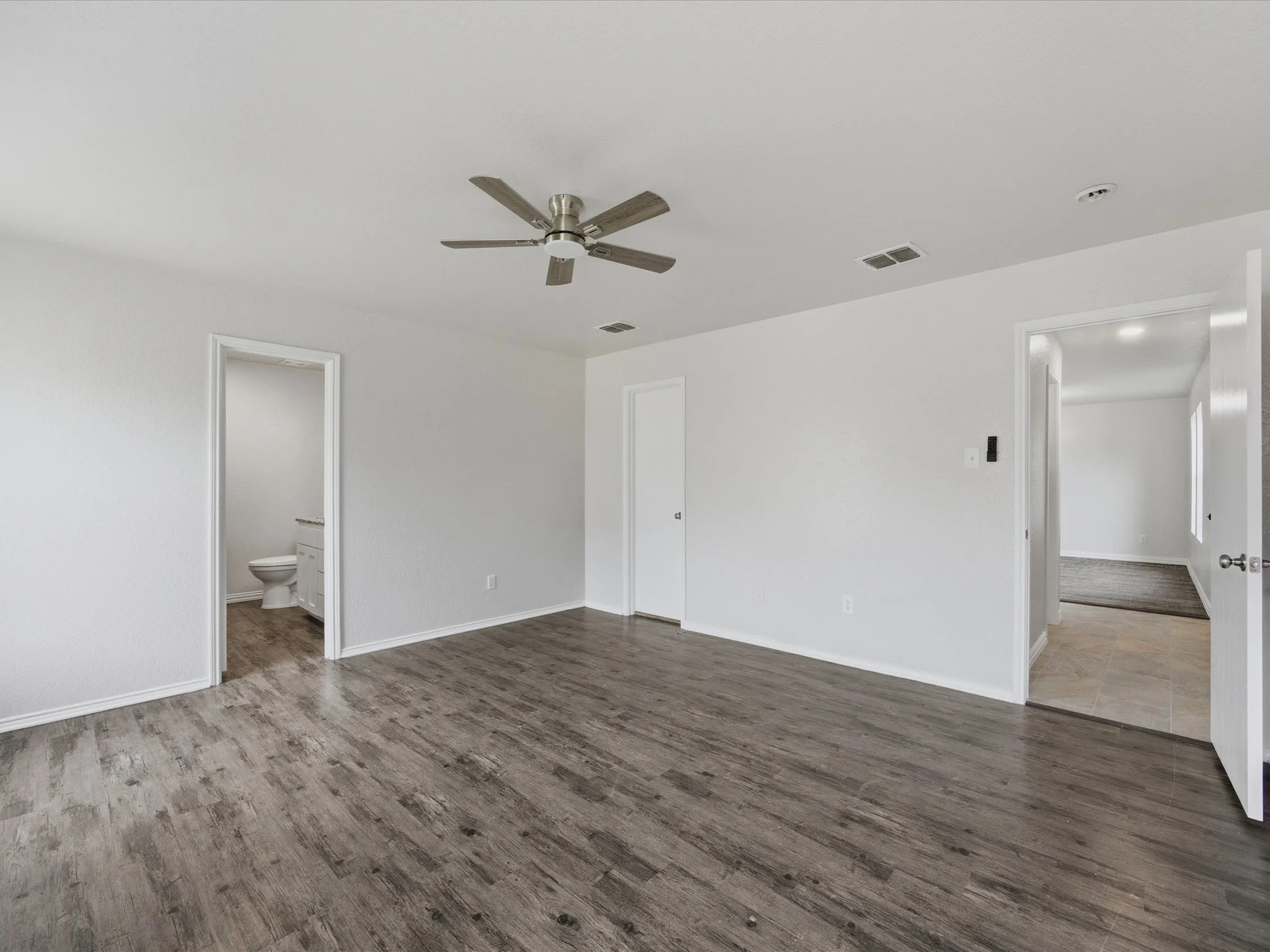 Empty living room with white walls, hardwood floors, a ceiling fan, and open doorways leading to other rooms including a bathroom.