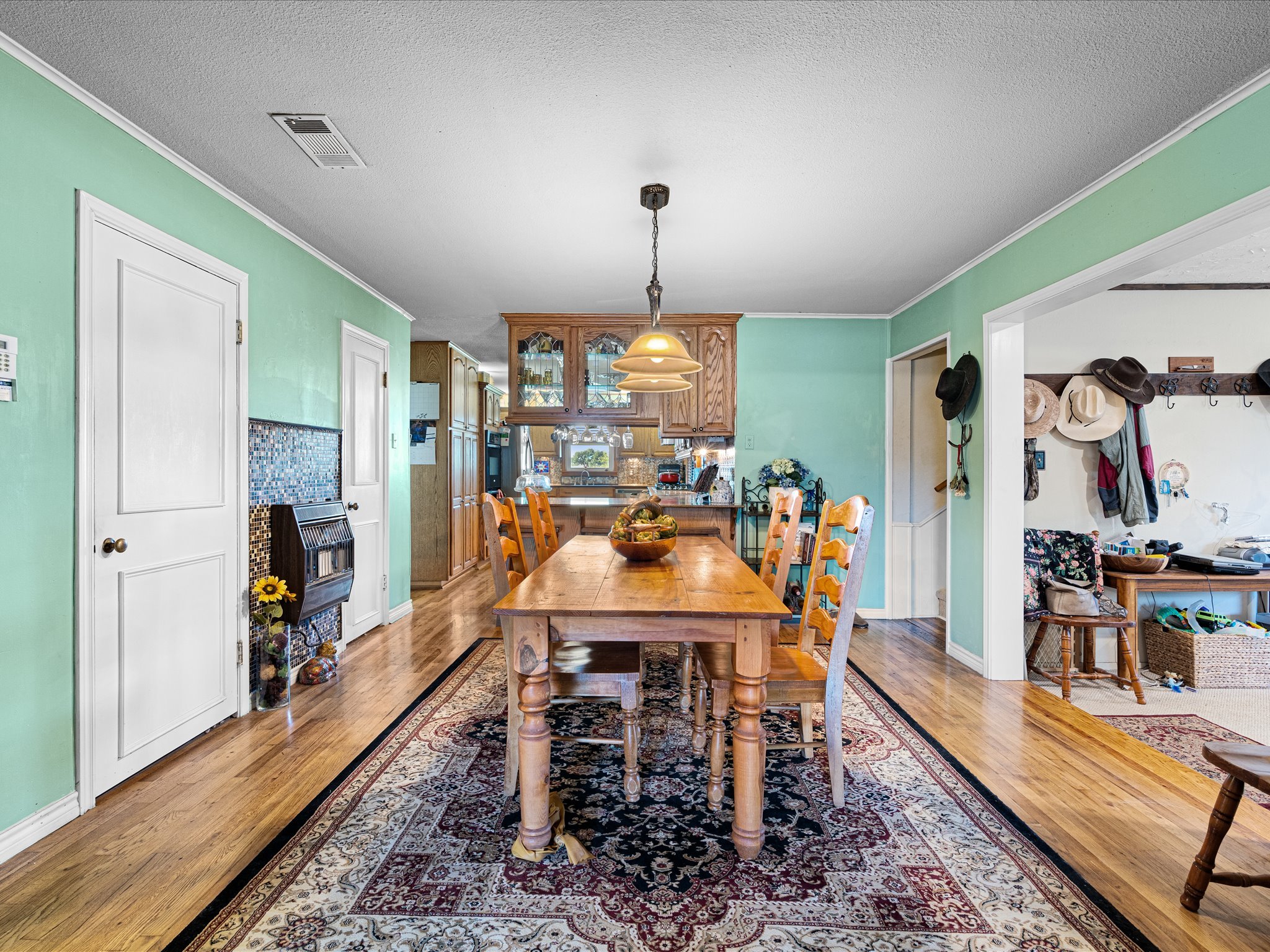 A dining room with a wooden table and matching chairs on a decorative area rug, with light green walls, a wooden cabinet, and a view into a kitchen area.
