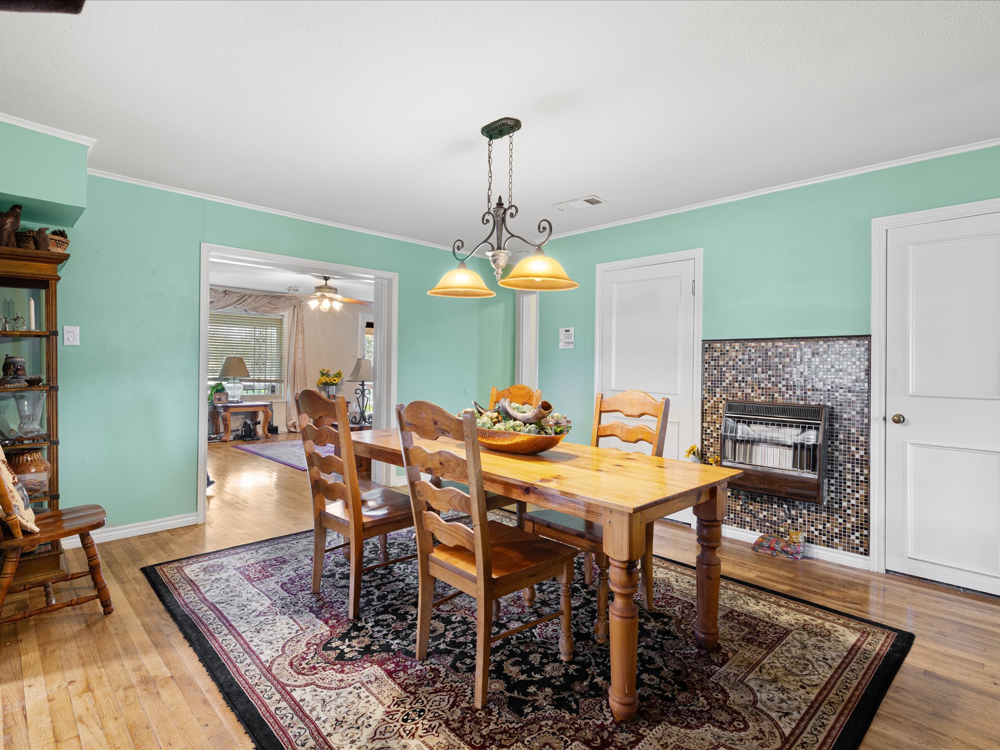 A dining room with a wooden table surrounded by matching chairs, a rug underneath, a decorative chandelier hanging from the ceiling, a fireplace with a mosaic tile design, and rooms visible through doorways leading to other areas of the house.