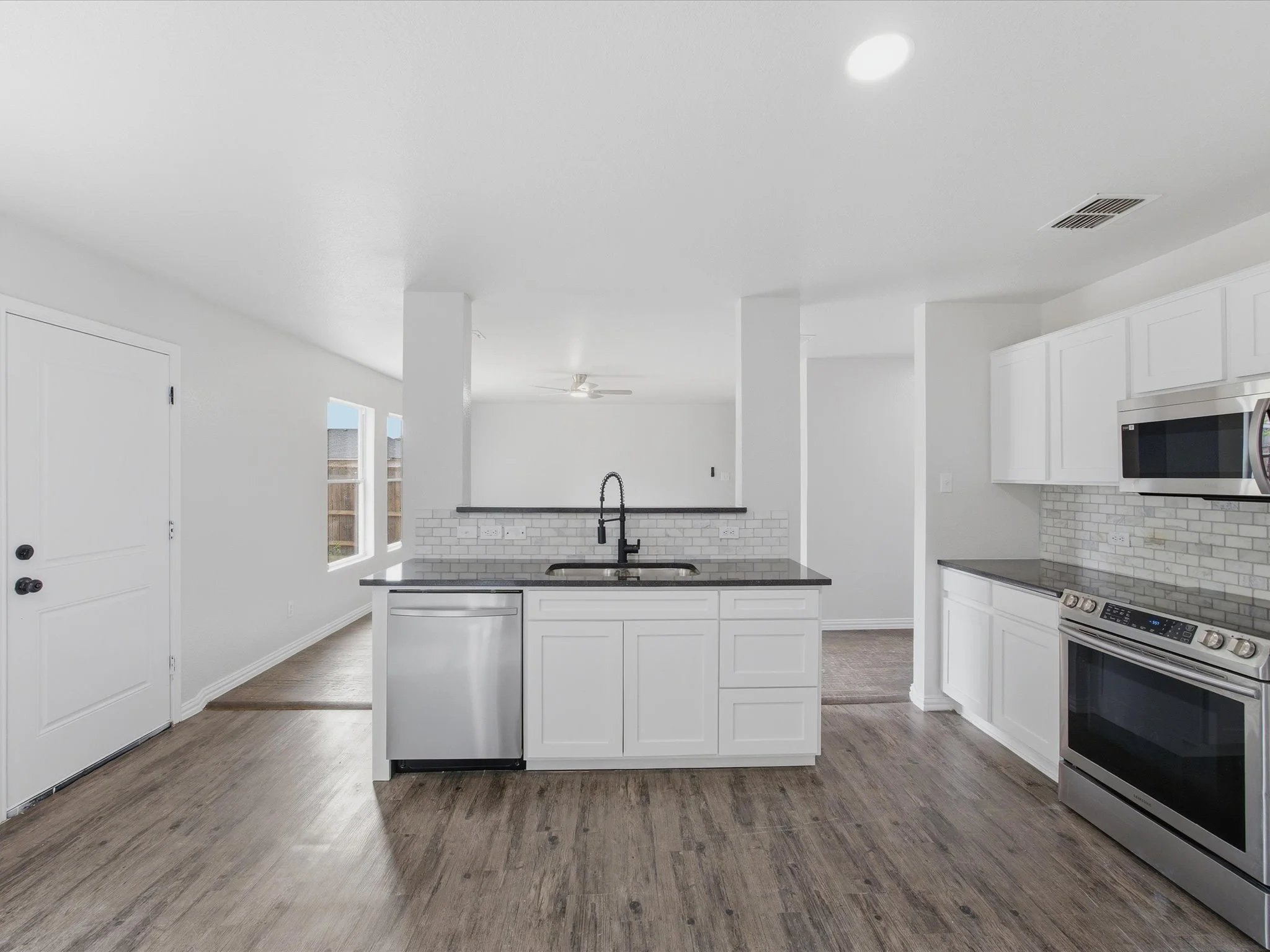 Modern kitchen with white cabinets, brick backsplash, black countertop island, stainless steel oven and microwave, hardwood floor, and large windows.