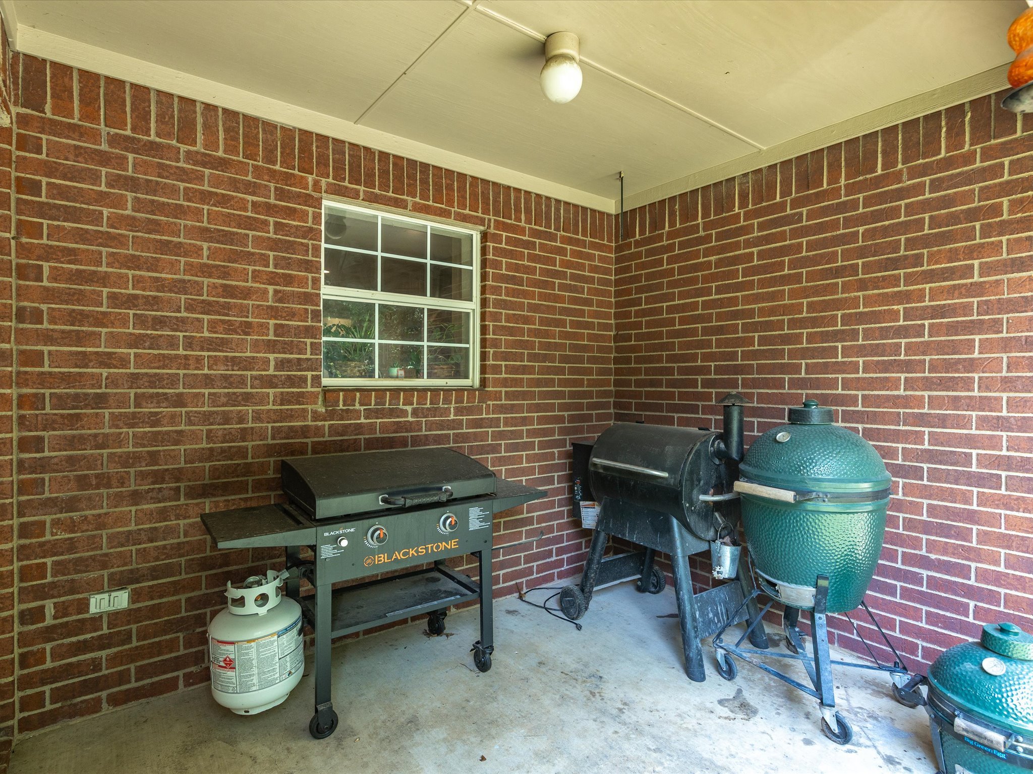 A covered patio area with brick walls, a small window, and outdoor cooking equipment including a Blackstone griddle, a smoker, and a green egg grill. There is also a propane tank and a power outlet on the wall.