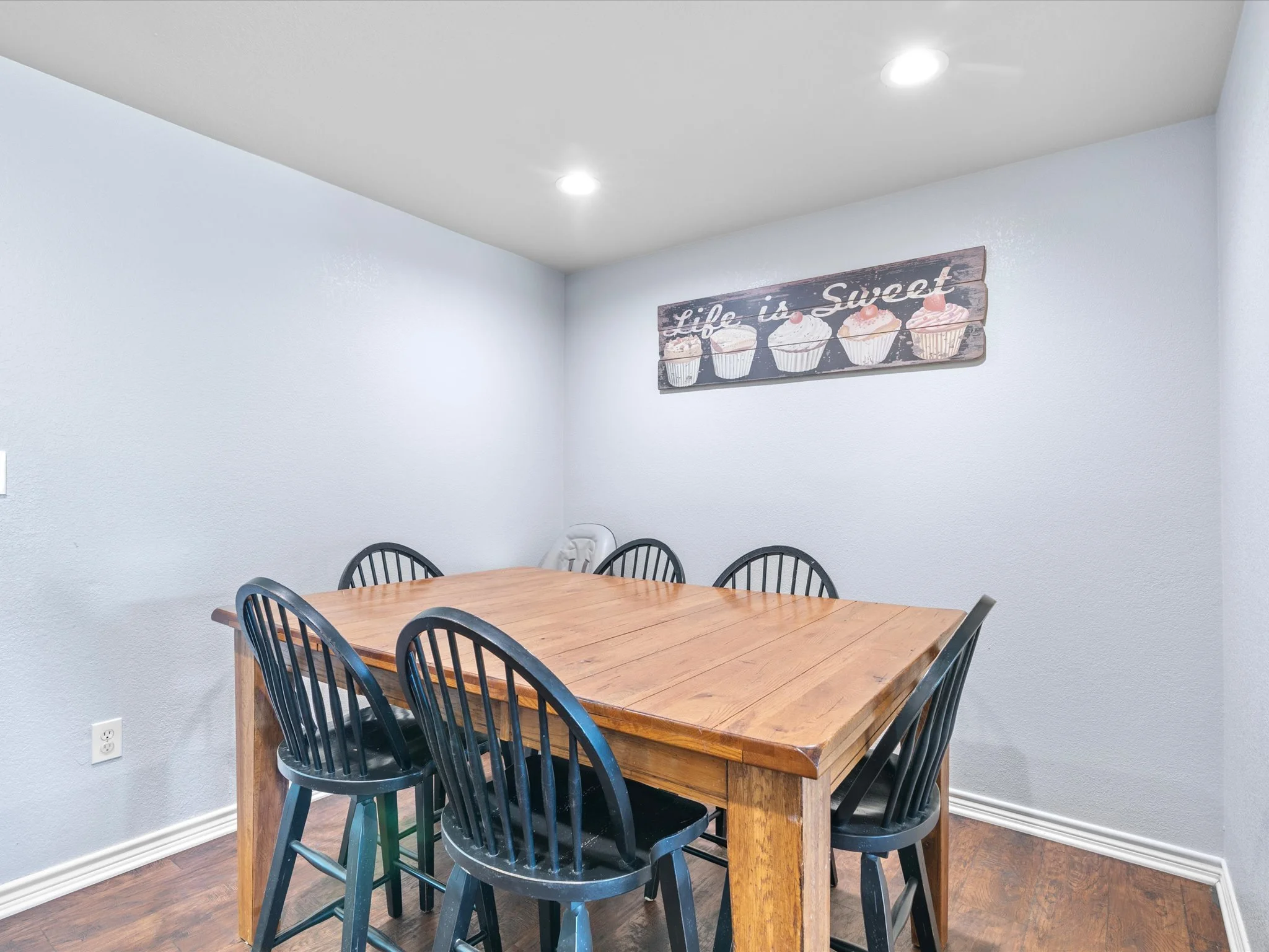 A wooden dining table surrounded by black chairs in a room with white walls, hardwood flooring, and a wall hanging that reads 'Life is Sweet' with images of cupcakes.