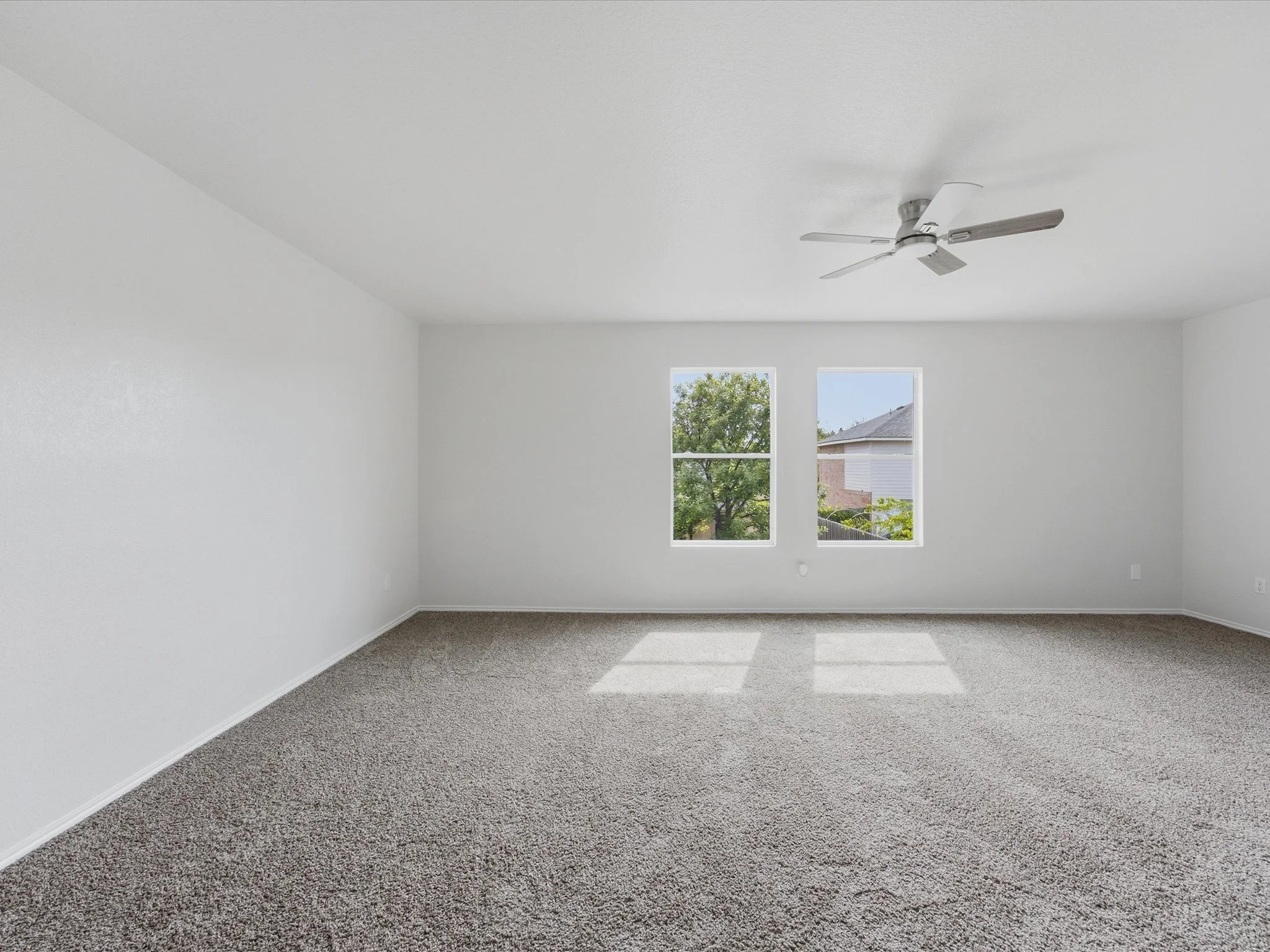 Empty room with white walls, two windows showing trees and a house outside, gray carpeted floor, and a ceiling fan.