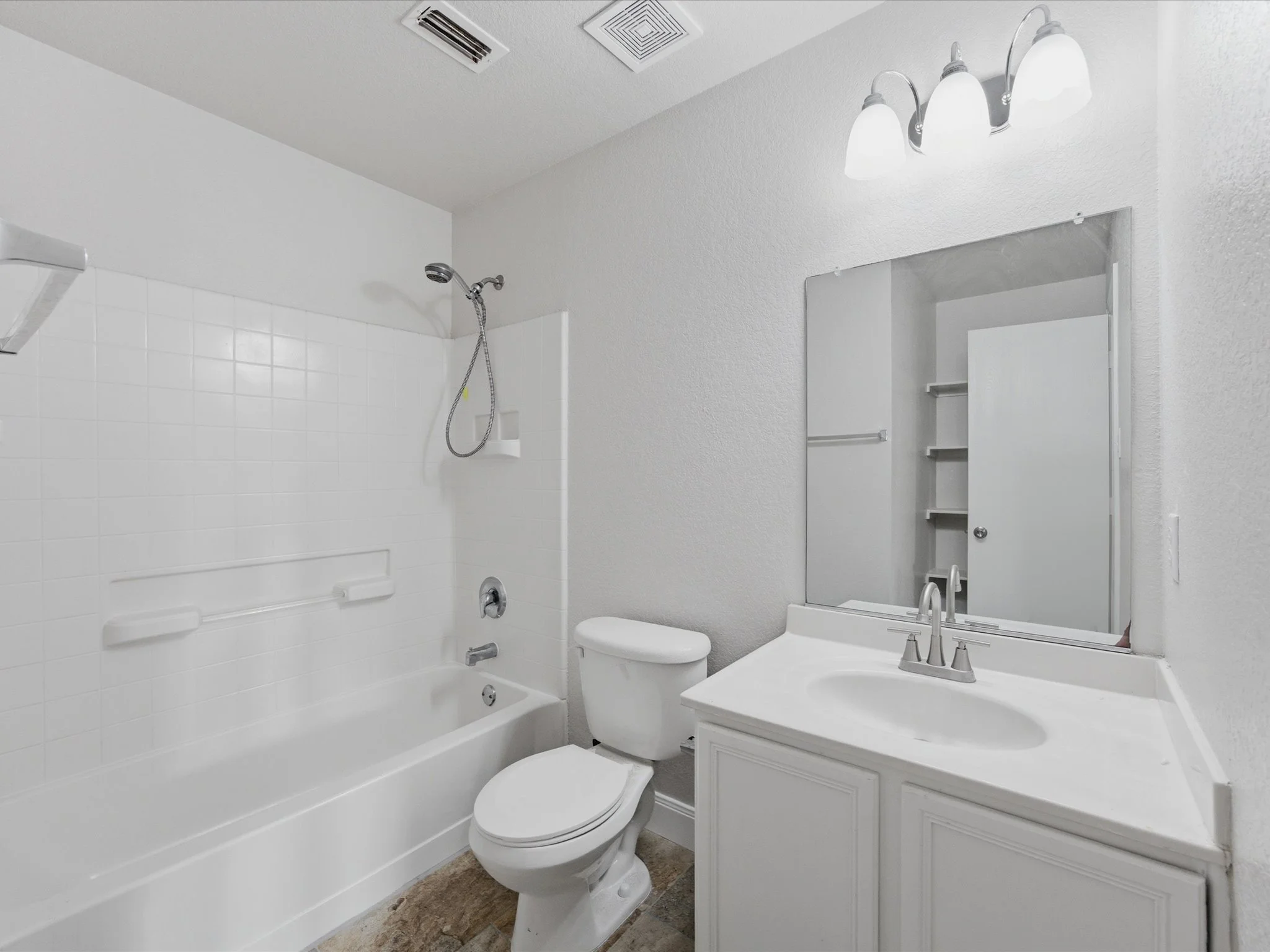 A small bathroom featuring a bathtub with a showerhead, white tiled walls, a toilet, a vanity with a sink, a mirror, and a three-light fixture above the mirror.