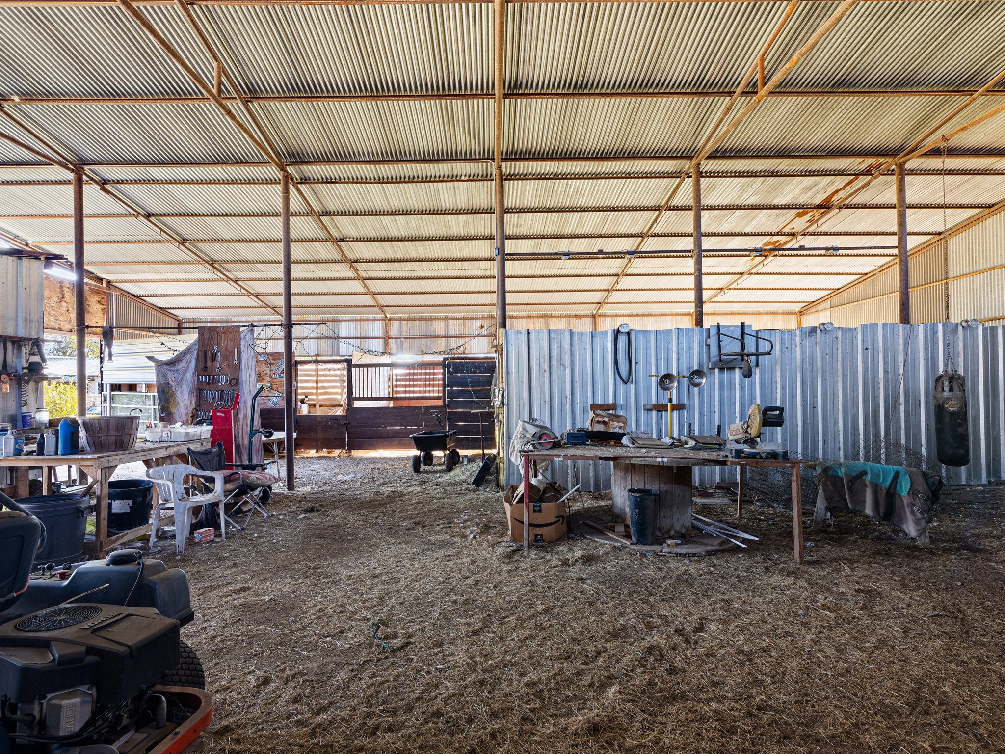 Interior view of a metal workshop or shed with a corrugated metal roof and walls. The space is cluttered with various tools, equipment, and containers, and has a dirt or straw-covered floor.