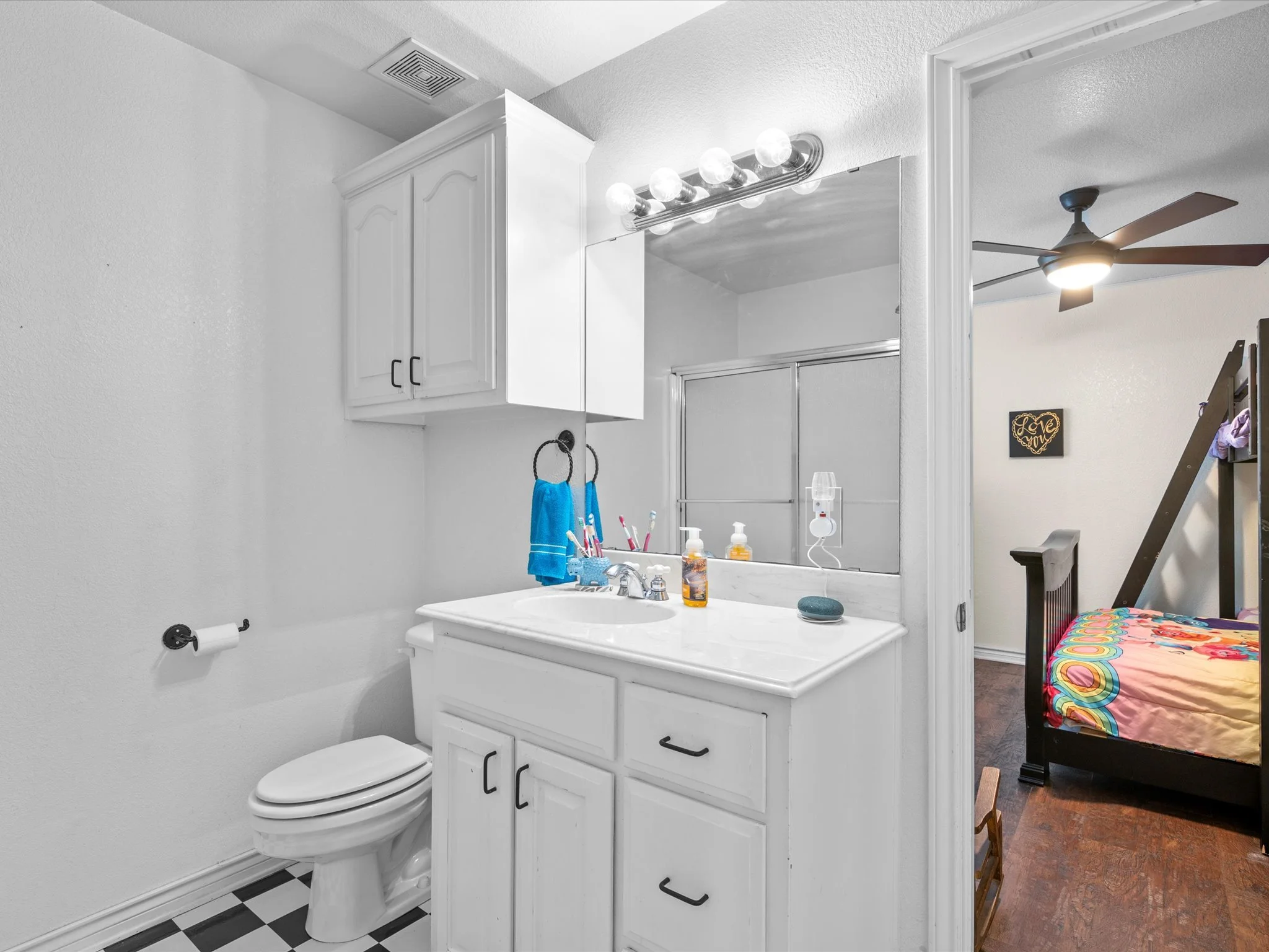 A bathroom with a white vanity and mirror, lit by four round bulbs, with a toothbrush and soap on the sink. To the right, an open doorway reveals a bedroom with a colorful bedspread and a ceiling fan, and a small wall decoration with the phrase 'Love