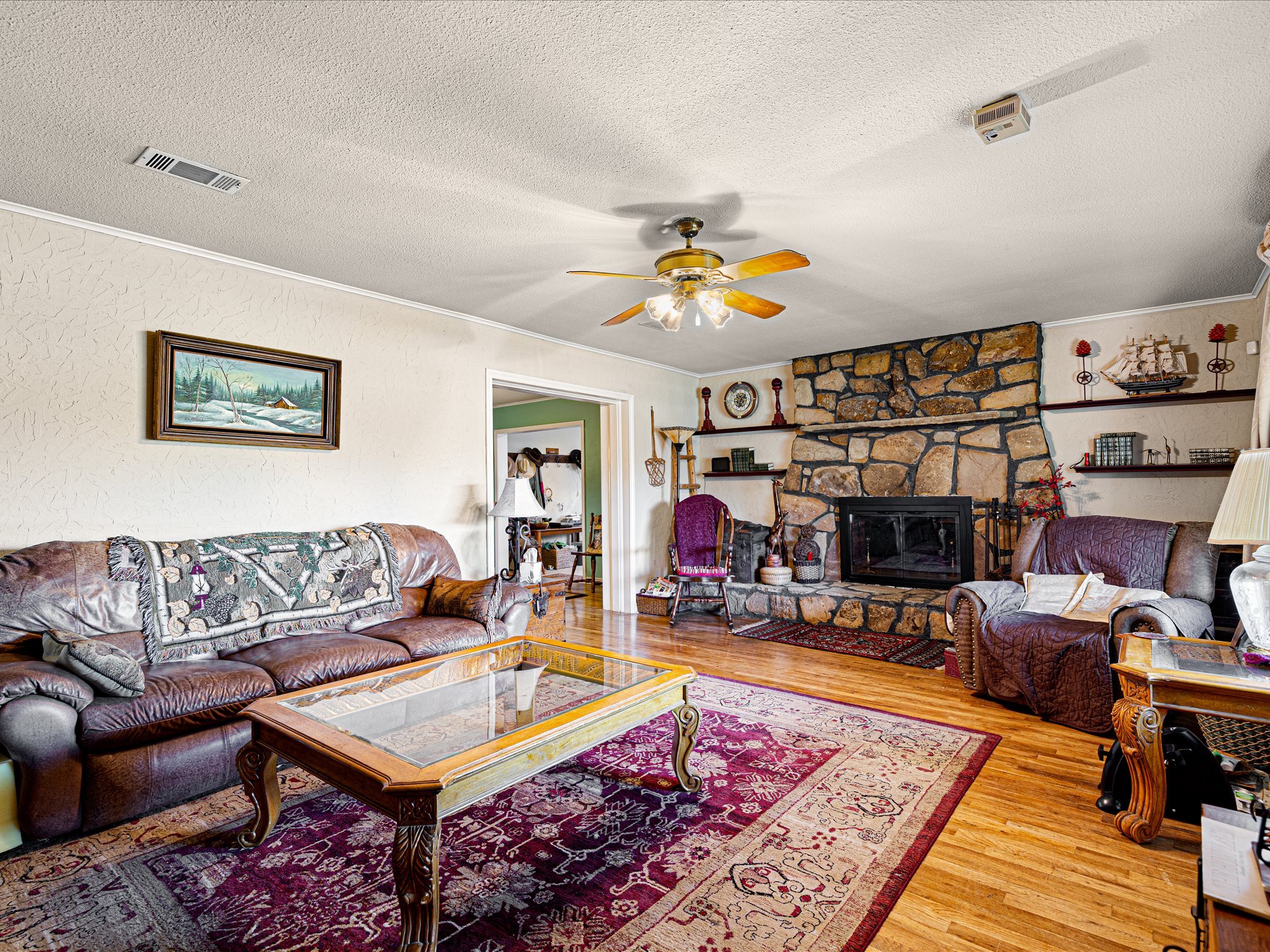 Living room with a leather sofa, a quilted armchair, a glass-topped coffee table, a red patterned area rug, a stone fireplace, wall art, a ceiling fan, and various decorative items.