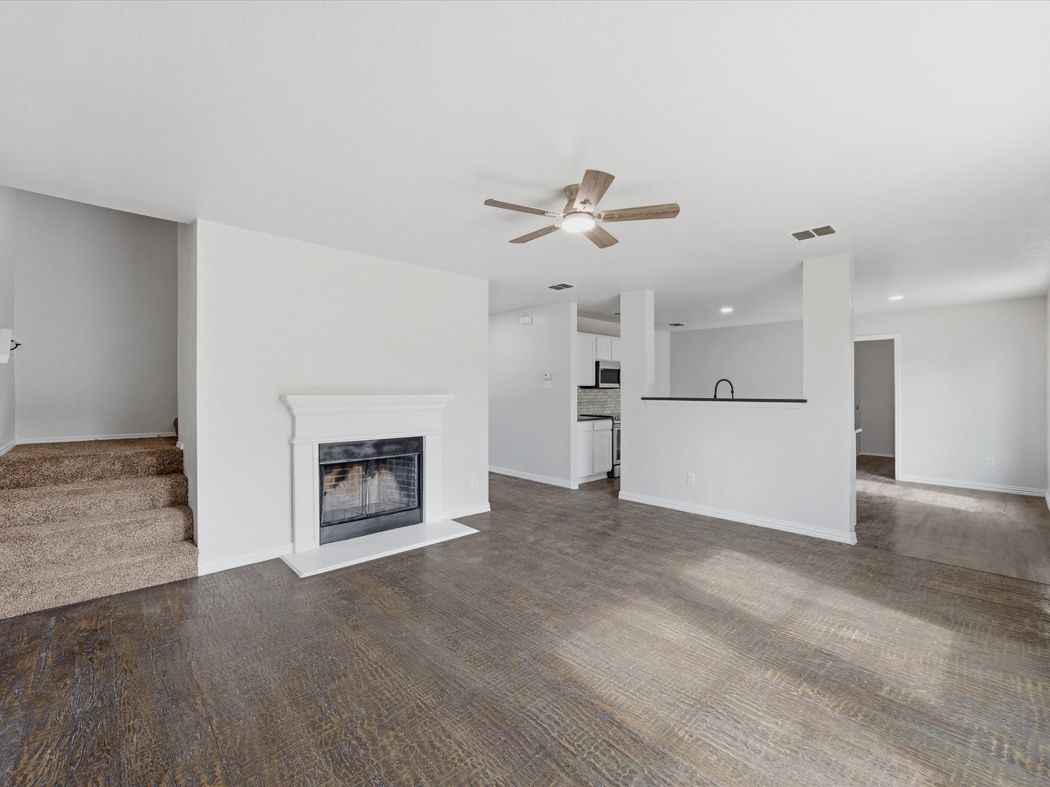 Interior view of a living room with a fireplace, ceiling fan, and open kitchen area with white cabinets and appliances. Carpeted stairs lead to other rooms.