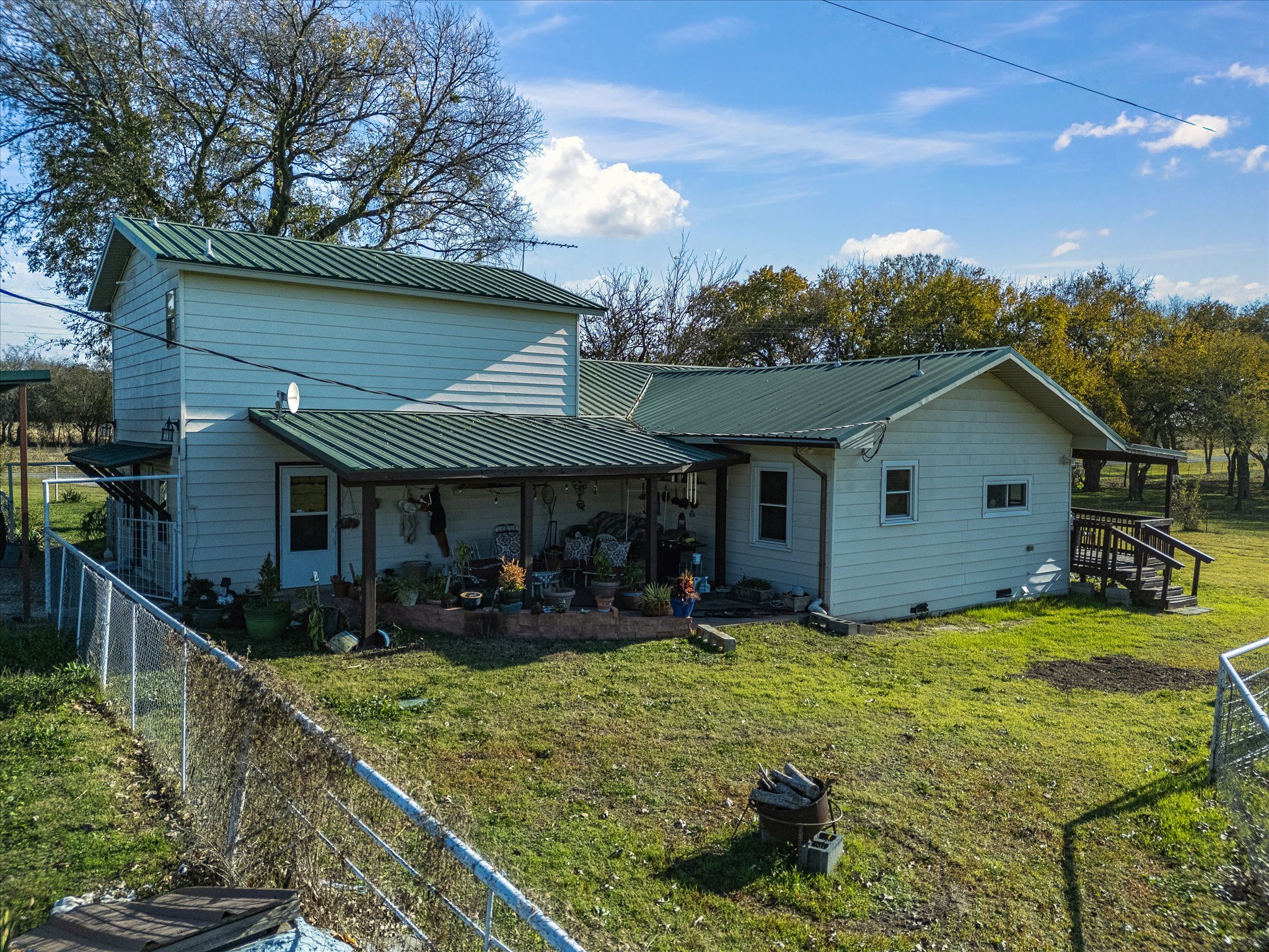 A two-story house with a green metal roof, a porch area with outdoor furniture, and a white exterior. Surrounding the house is a grassy yard enclosed by a chain-link fence. In the background, there are trees with some leaves changing color and a part