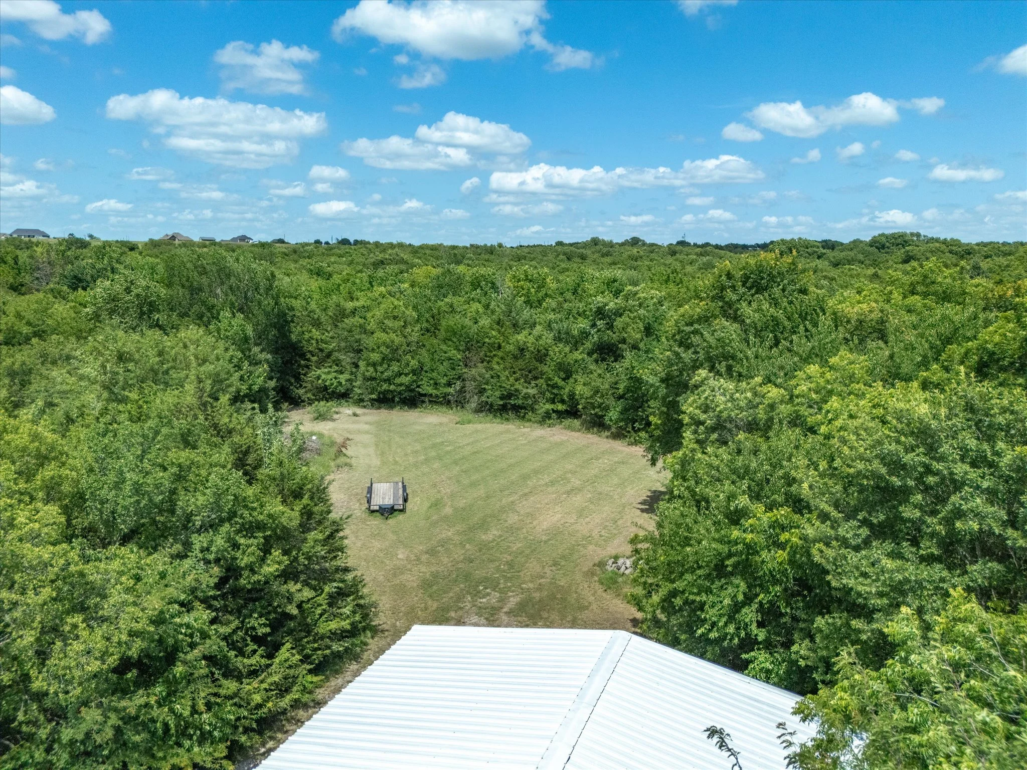 A lush green landscape with trees, a field, and a metal roof in the foreground. Blue sky with scattered clouds overhead.