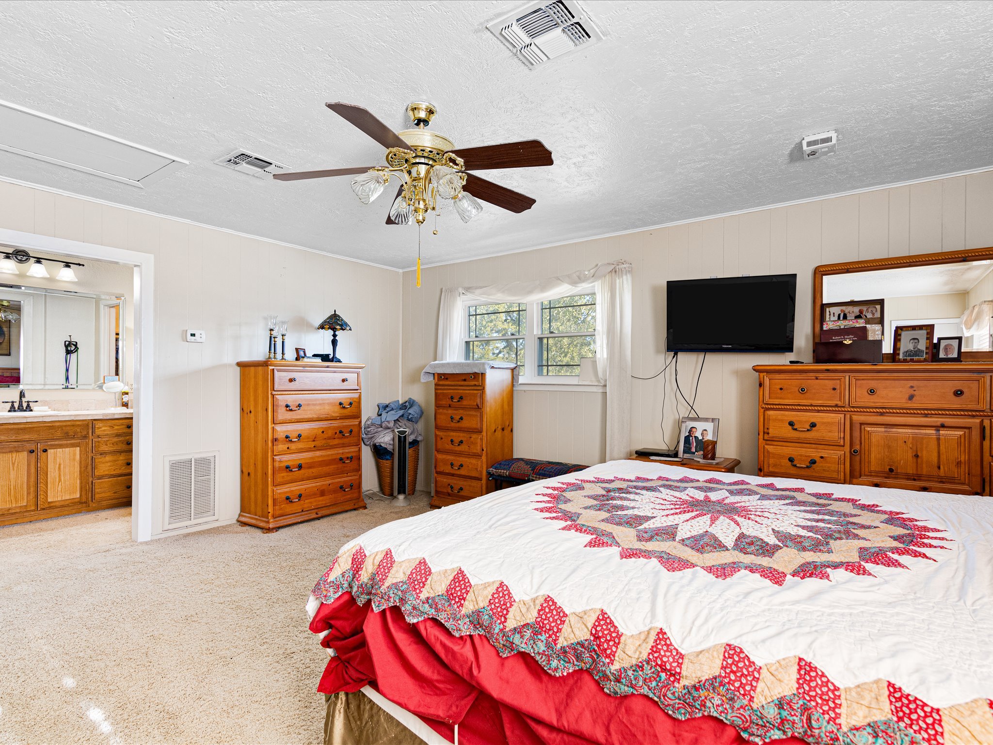 A bedroom with a bed covered by a colorful quilt, wooden dressers, a window with curtains, a bedside table with photos, a wall-mounted TV, and a ceiling fan.