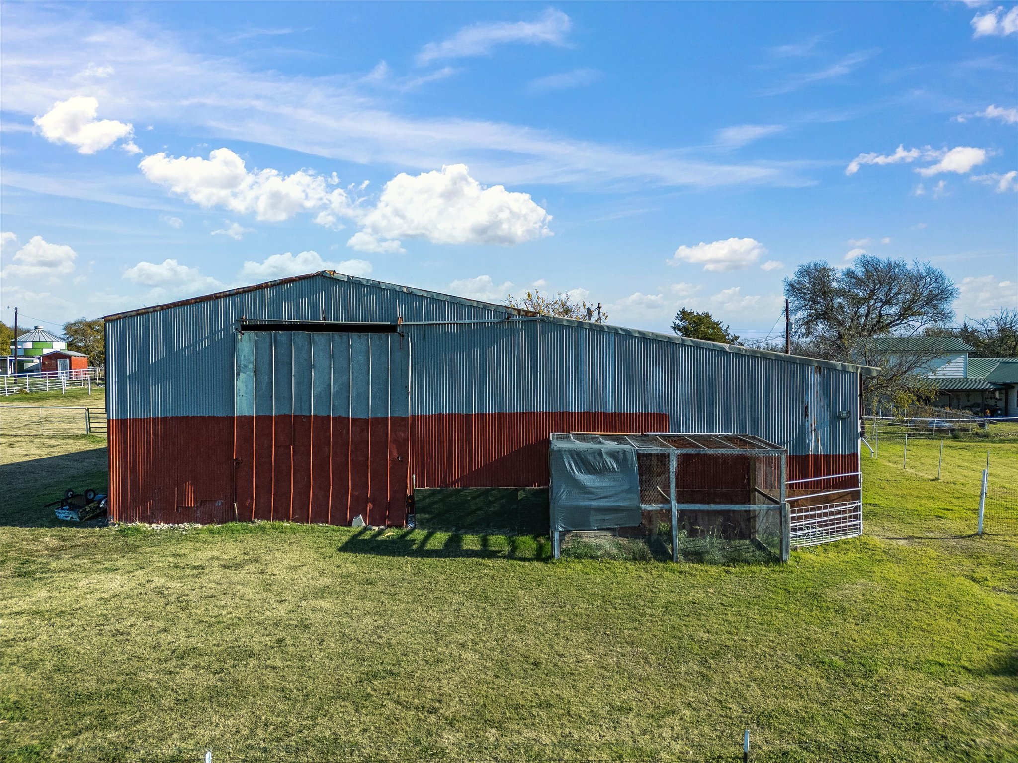 A large blue and red metal barn with some trees and houses in the background under a partly cloudy sky.