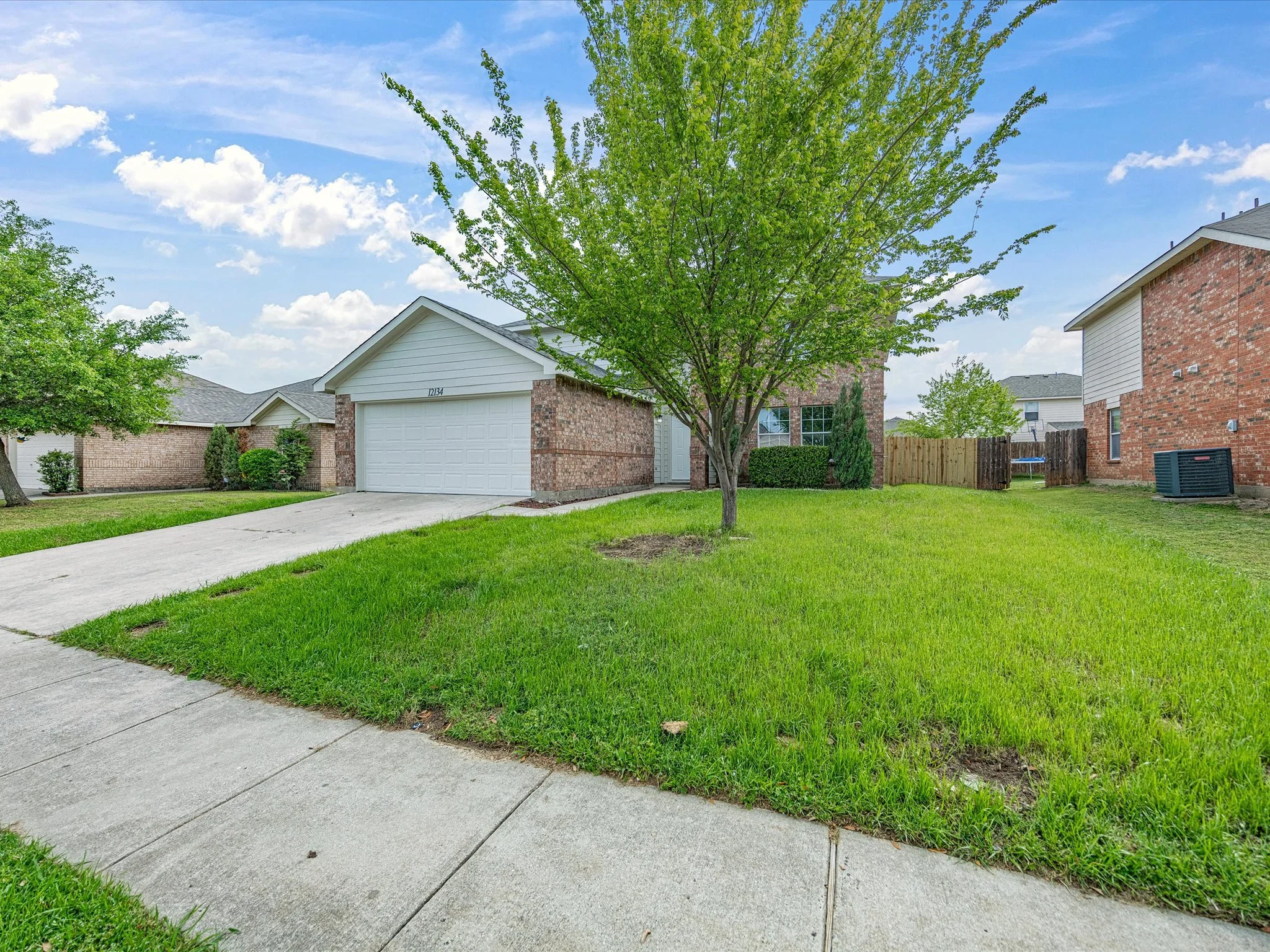 A suburban house with a brick exterior, white garage door, and a gabled roof, surrounded by a green lawn and trees under a partly cloudy sky.