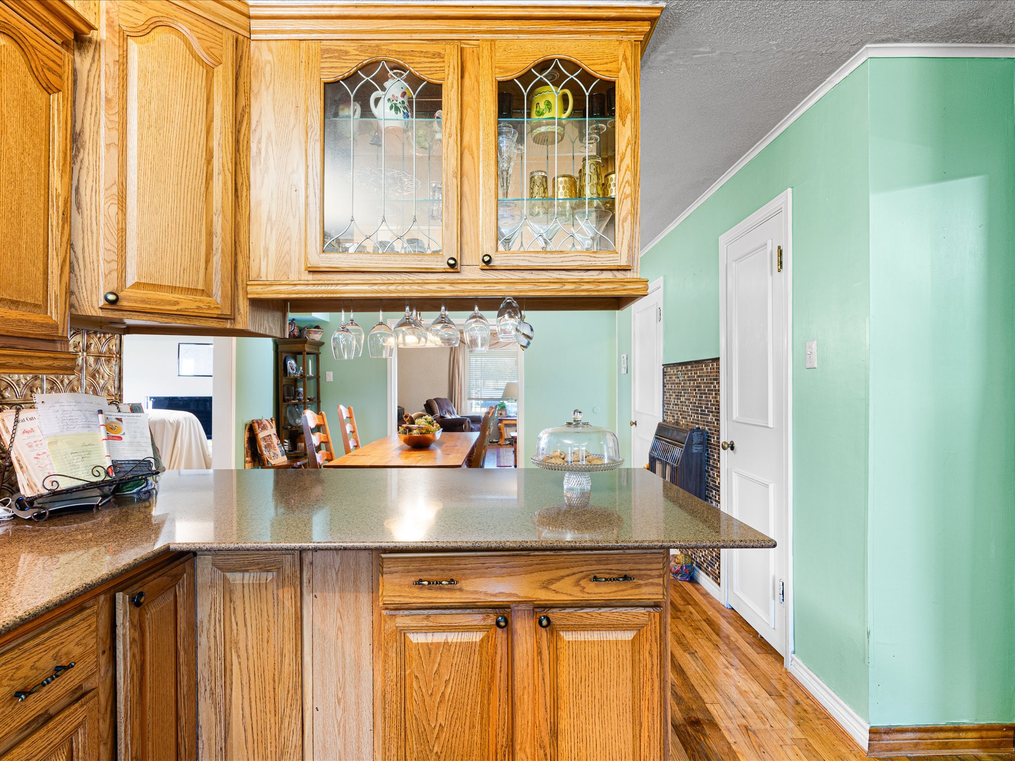 View of a kitchen with wooden cabinets, a block of granite countertop, a green painted wall, a white door and a dining area with wooden chairs and a table, with a refrigerator in the background.