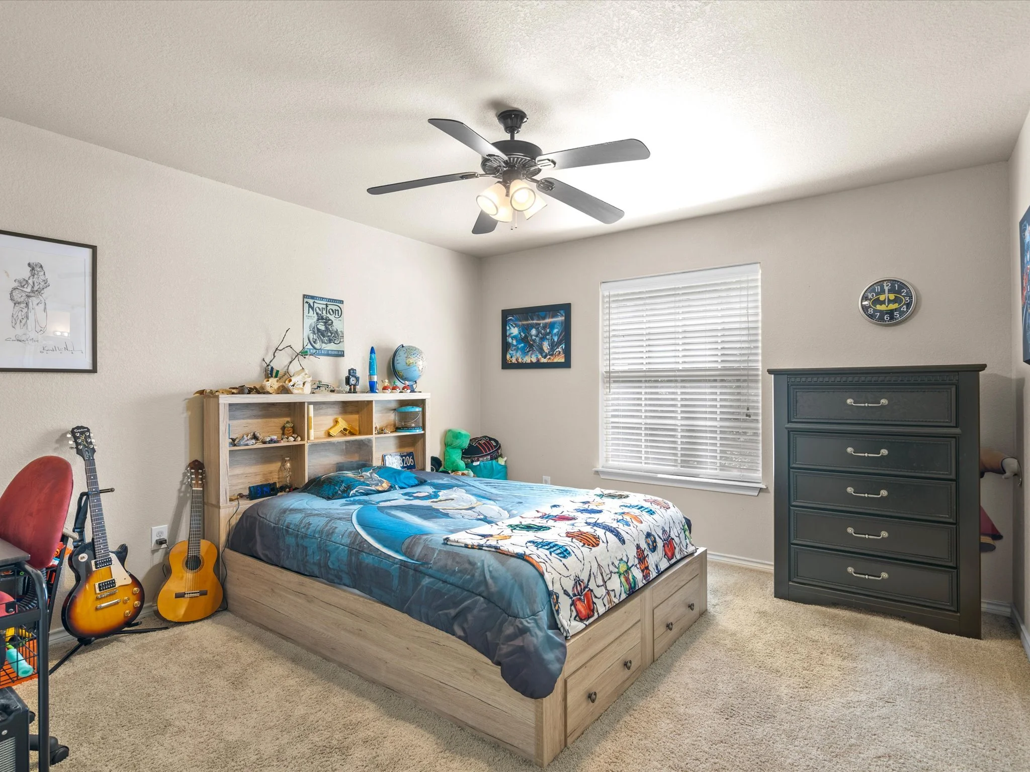 A bedroom with a wooden bed, colorful bedspread, and a gray dresser. Two guitars are leaning against the wall, and various sports memorabilia and decorations are on a bookshelf. There are framed posters and a clock on the walls, with a window covered