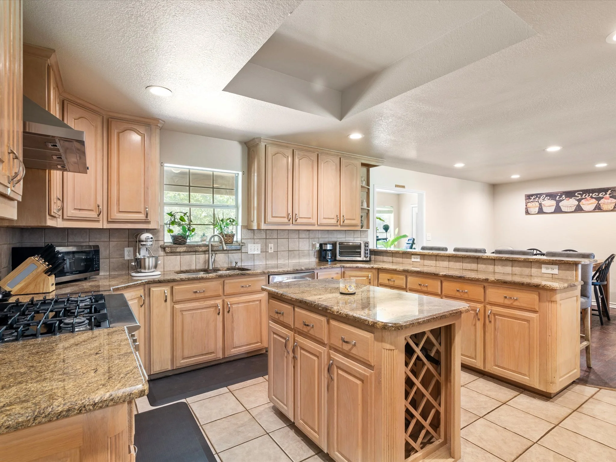 A spacious kitchen with light wood cabinets, a central island with a granite countertop, a built-in wine rack, and stainless steel appliances. There is a window above the sink, potted plants on the windowsill, and a banner that reads 'Life is Sweet' 