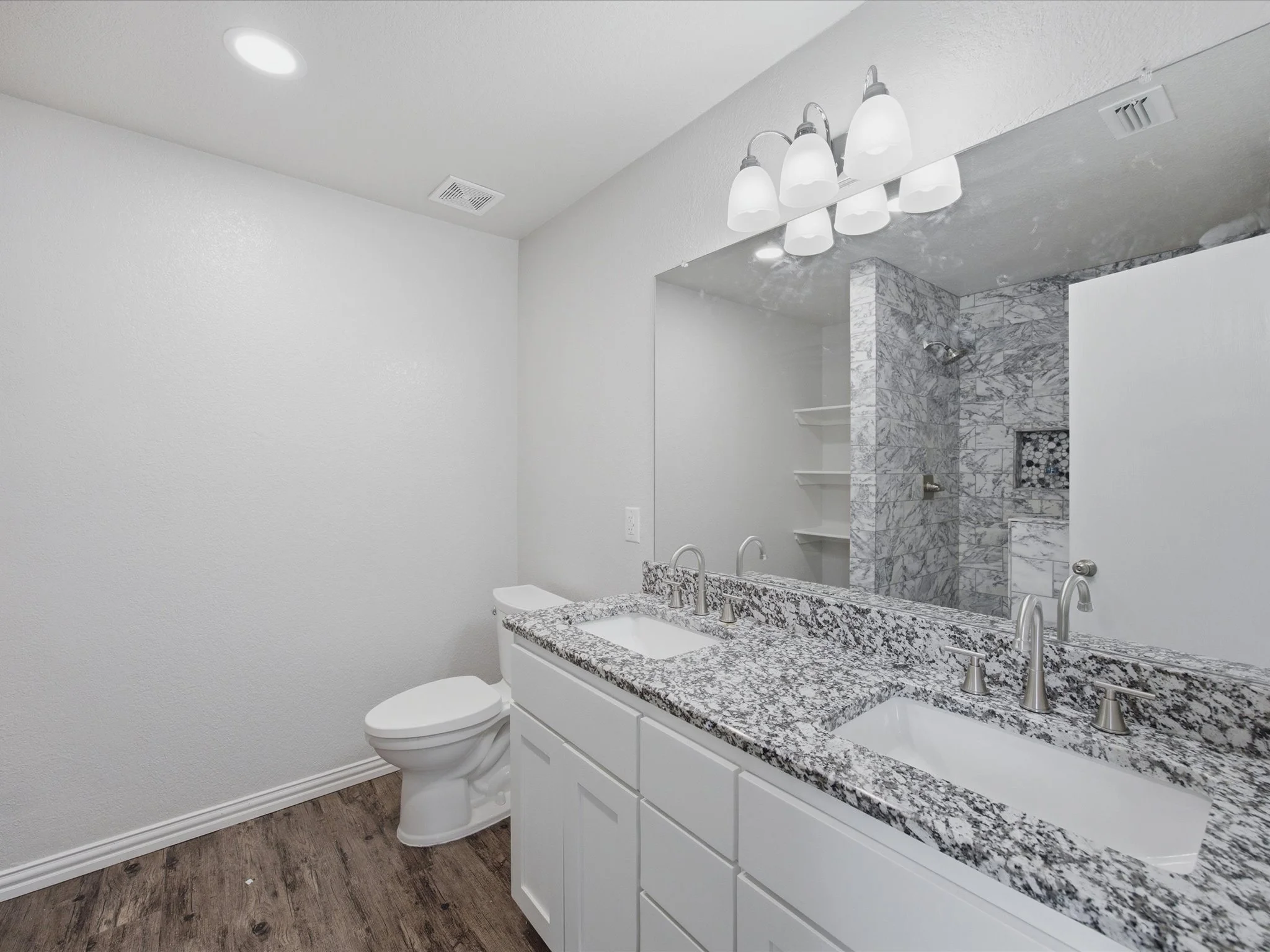 Modern bathroom with a white vanity, granite countertop, large mirror, and toilet. The shower area features gray marble tiles, a built-in shelf, and a rainfall showerhead. The ceiling has recessed lighting and an exhaust vent. The floor is wood-look 