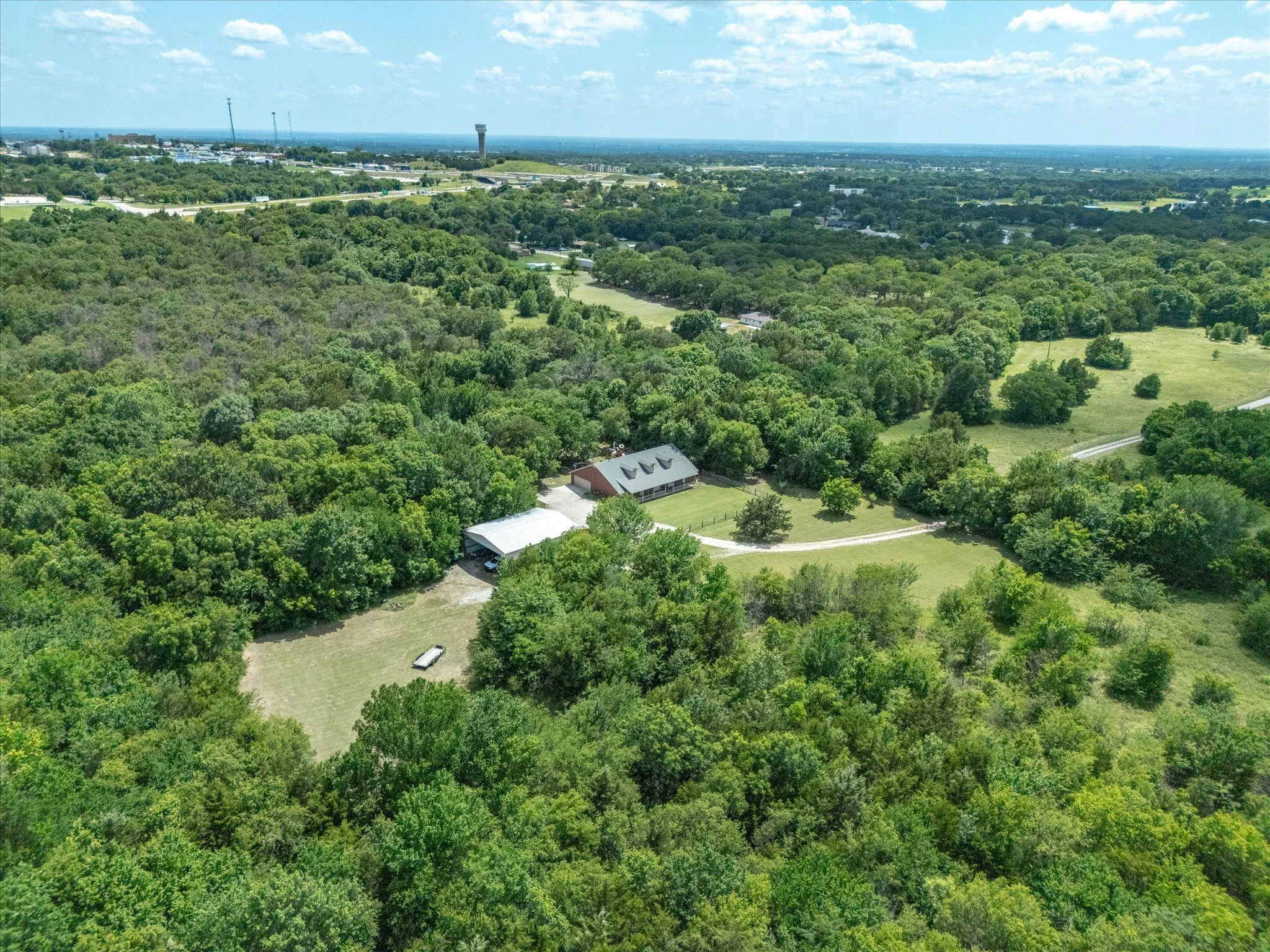 Aerial view of a lush green landscape with trees, open fields, and a few houses. In the distance, there are roads, a water tower, and a city skyline under a partly cloudy sky.