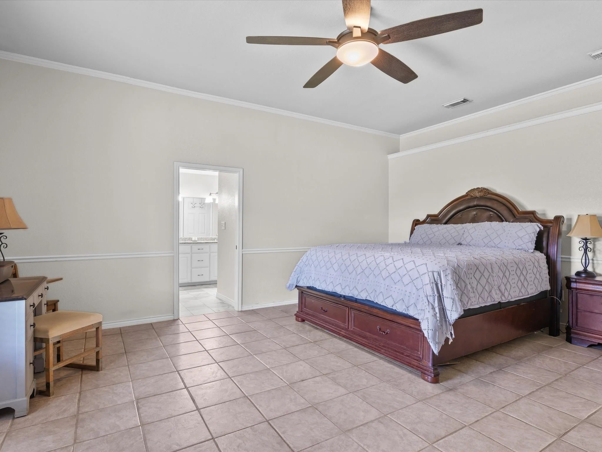 A bedroom with beige walls, a wooden bed with a white patterned bedspread, and nightstands with lamps. There is a ceiling fan with wooden blades and a light fixture. An opening shows a bathroom with white cabinets and a countertop.